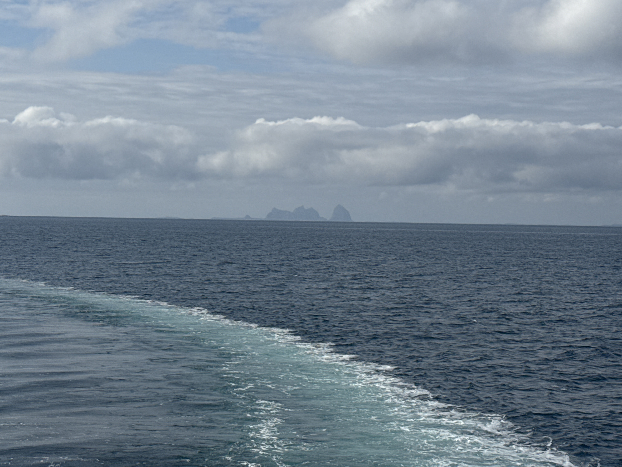This image depicts a wide, calm, deep blue ocean under a cloudy sky with distant, hazy islands. In the foreground, a white foamy wake trails from the left side of the frame, indicating movement through the water, likely from a boat. The horizon is visible, separating the sea from the sky, where numerous grey and white clouds are scattered. The distant islands appear as dark, silhouetted shapes against the hazy background, suggesting a considerable distance from the viewpoint. The overall scene is one of open water and overcast weather.