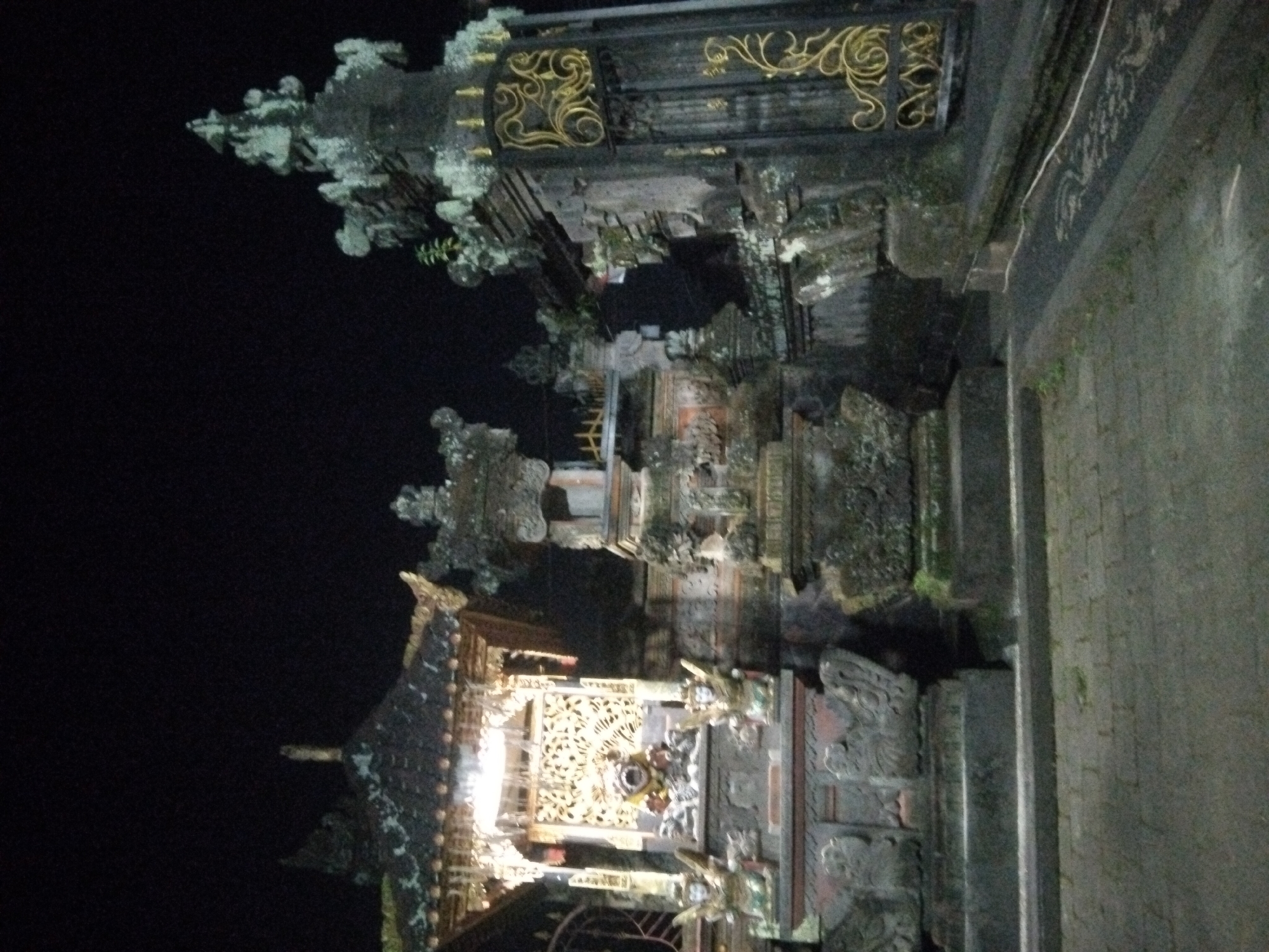 The image depicts a Balinese Hindu temple entrance at night. The entrance features ornate stone carvings with gold accents and tiered roofs. The surrounding environment is dark, with the temple's features highlighted by artificial lighting. The location is identified as Unknown, Unknown.