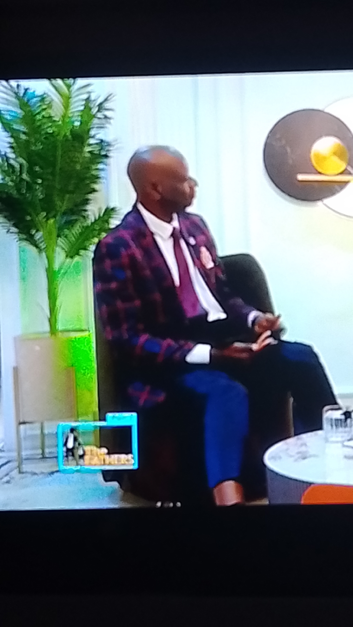 A man with dark skin and a bald head is seated in a chair, wearing a plaid suit jacket with purple and blue checkered patterns, a white collared shirt, and a purple tie. He is looking to his right. To his left is a tall, potted plant with green, feathery leaves in a light-colored vase with a green hue. In the lower left corner of the image, there is a small blue-bordered rectangle displaying a logo with the words "THE FATHERS" in gold lettering. In the background, there is a circular wall decoration with a gold element. To the right of the man, a portion of a white table with an orange base is visible, with a clear glass object on it. The scene appears to be indoors, possibly in a studio setting, and there is no indication of it being located in Lokoja, Nigeria.