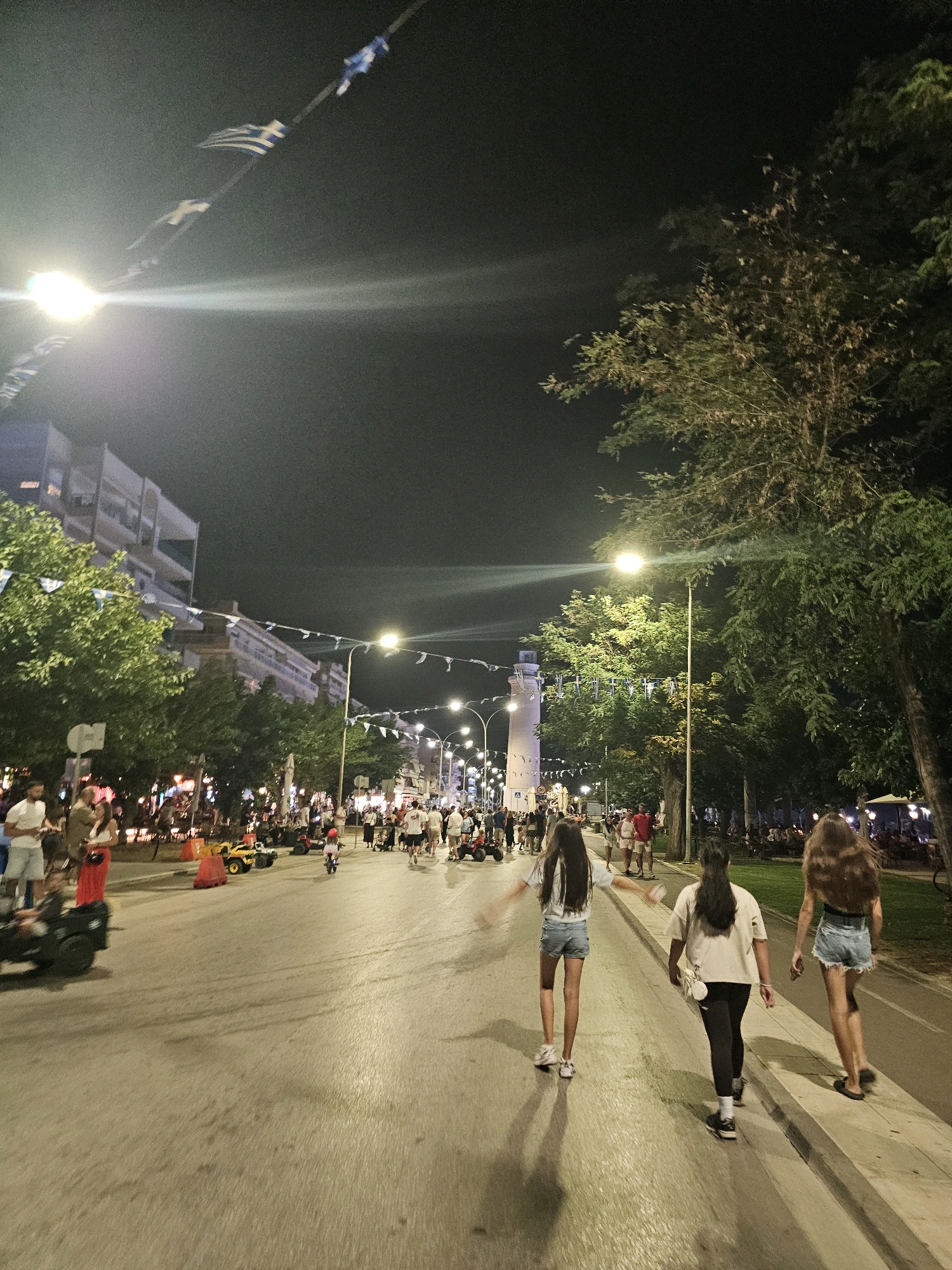 The image captures a night scene along a wide, paved street in Alexandroupolis, Greece. The street is lined with trees and buildings, and illuminated by streetlights and building lights. Greek flags are strung across the street, suggesting a festive or patriotic occasion. In the background, a lighthouse structure is visible. A diverse crowd of people are walking along the street and sidewalk. Some are casually dressed and appear to be enjoying an evening out. Children are seen on small ride-on vehicles. The atmosphere seems lively and social.