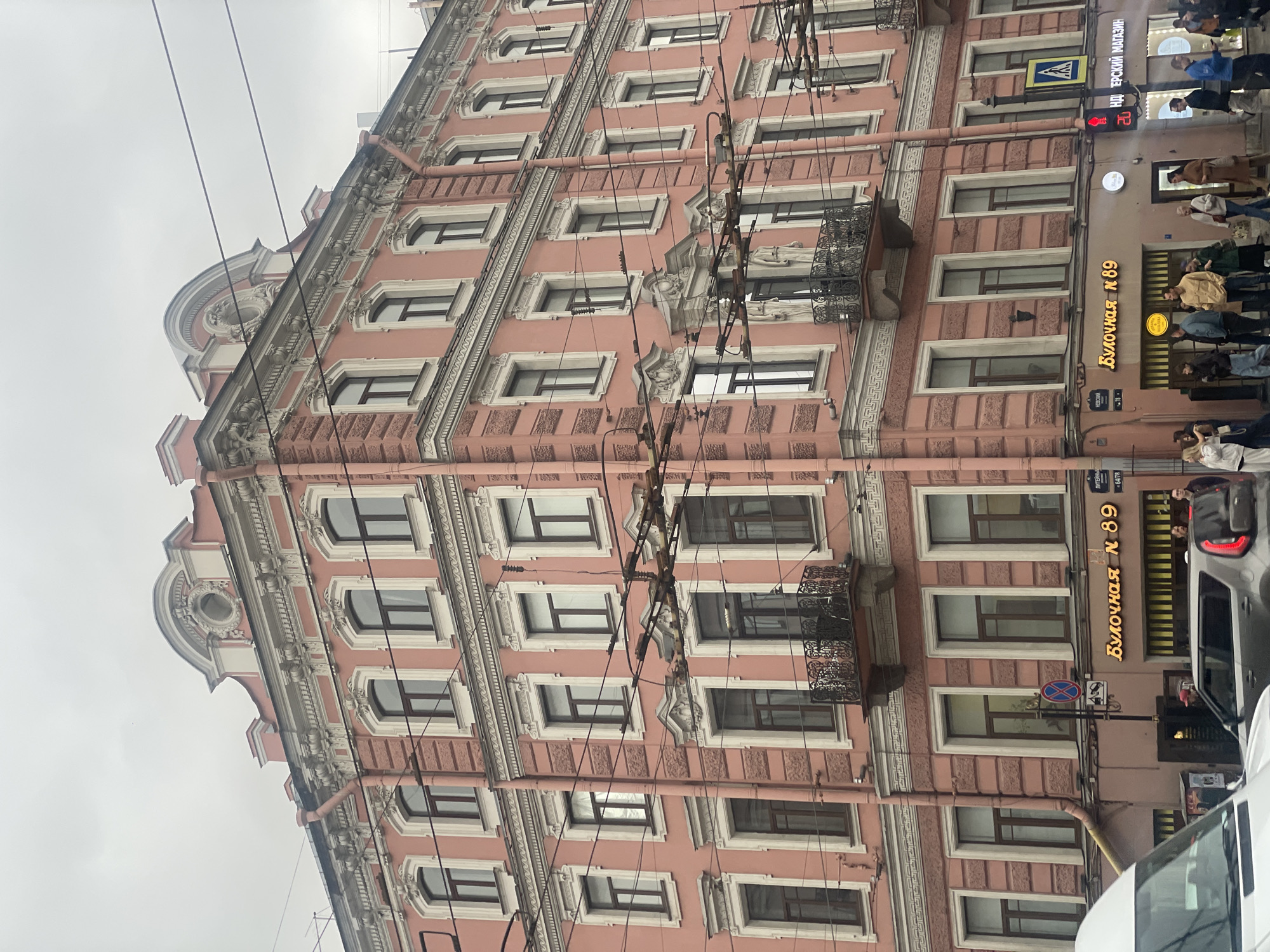The image shows a view of a pinkish building in Saint Petersburg, Russia. The building features multiple windows and ornate detailing on its facade. Electrical wires crisscross in front of the building. Signs are visible on the building, one illuminated with the words "Булочная № 89" (Bakery No. 89). People are walking on the sidewalk in front of the building. Part of a car is visible at the bottom right. A street sign with Russian text can be seen on the corner. A pedestrian crossing sign is positioned on a post. The sky is overcast.