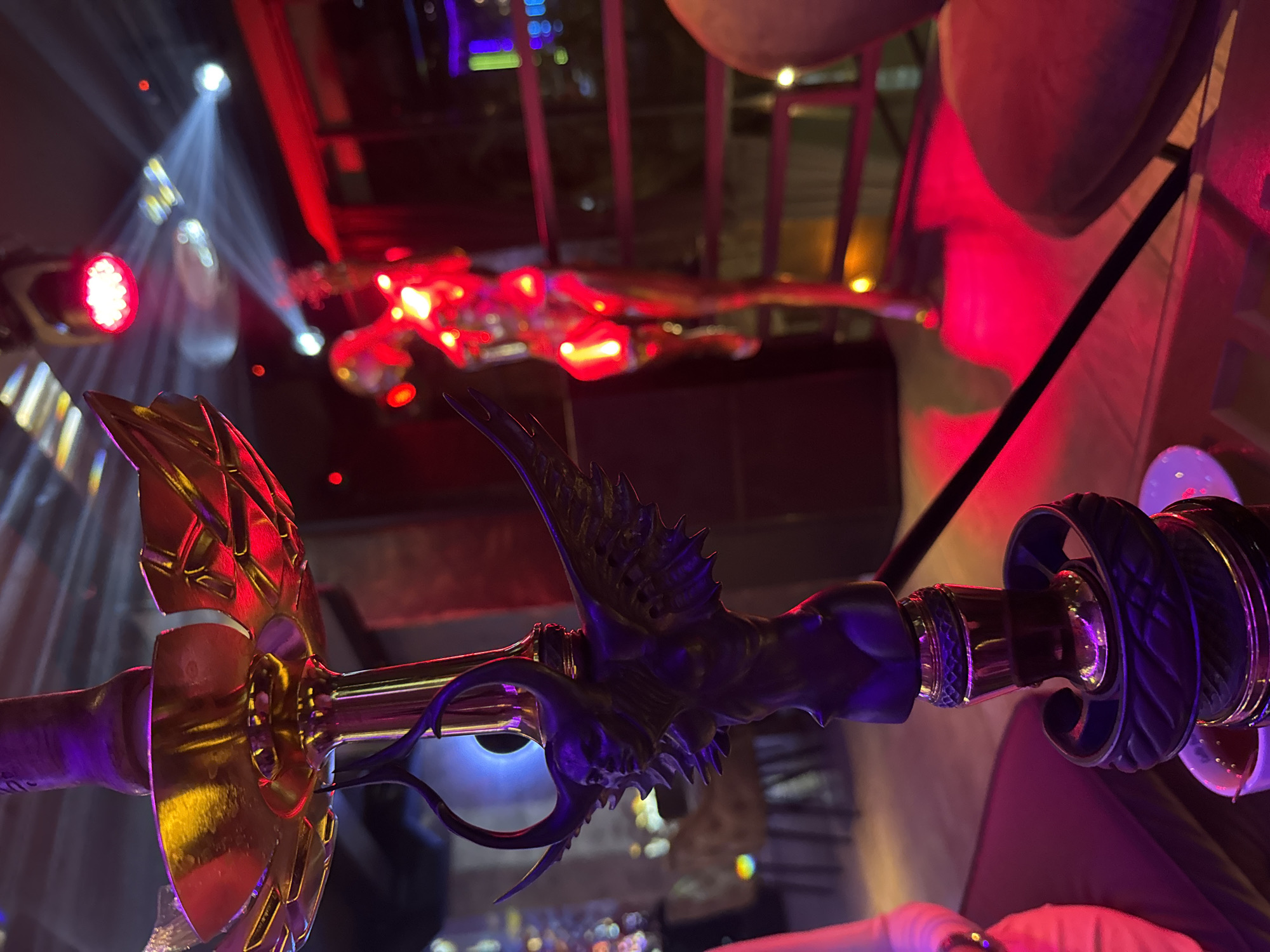 The image presents a stylized metal object, resembling a decorative weapon, under strong colored lighting. The object has elements of both gold and dark metal, with ornate detailing suggestive of a fantasy or historical design. The background includes out-of-focus lights and structures, possibly indicating an urban setting at night. The presence of colored stage lighting suggests this could be a bar, entertainment venue, or event location. Given the mention of Burgas, Bulgaria, this object and setting might be found within a themed restaurant, bar, or entertainment complex in the city.