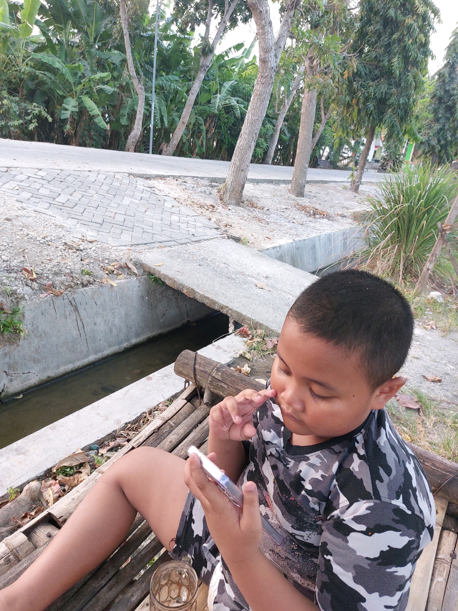 In Canga'an, Indonesia, a young boy with dark hair and fair skin is captured in a close-up shot. He is seated outdoors on a bamboo structure, wearing a camouflage t-shirt. The boy is looking down at a smartphone in his hands, with his right hand holding the device and his left hand touching his nose. Next to him, a glass with a condensation-covered interior rests on the bamboo. Behind him, a concrete drain runs alongside a path paved with interlocking stones, and lush green banana trees and other foliage form the backdrop.