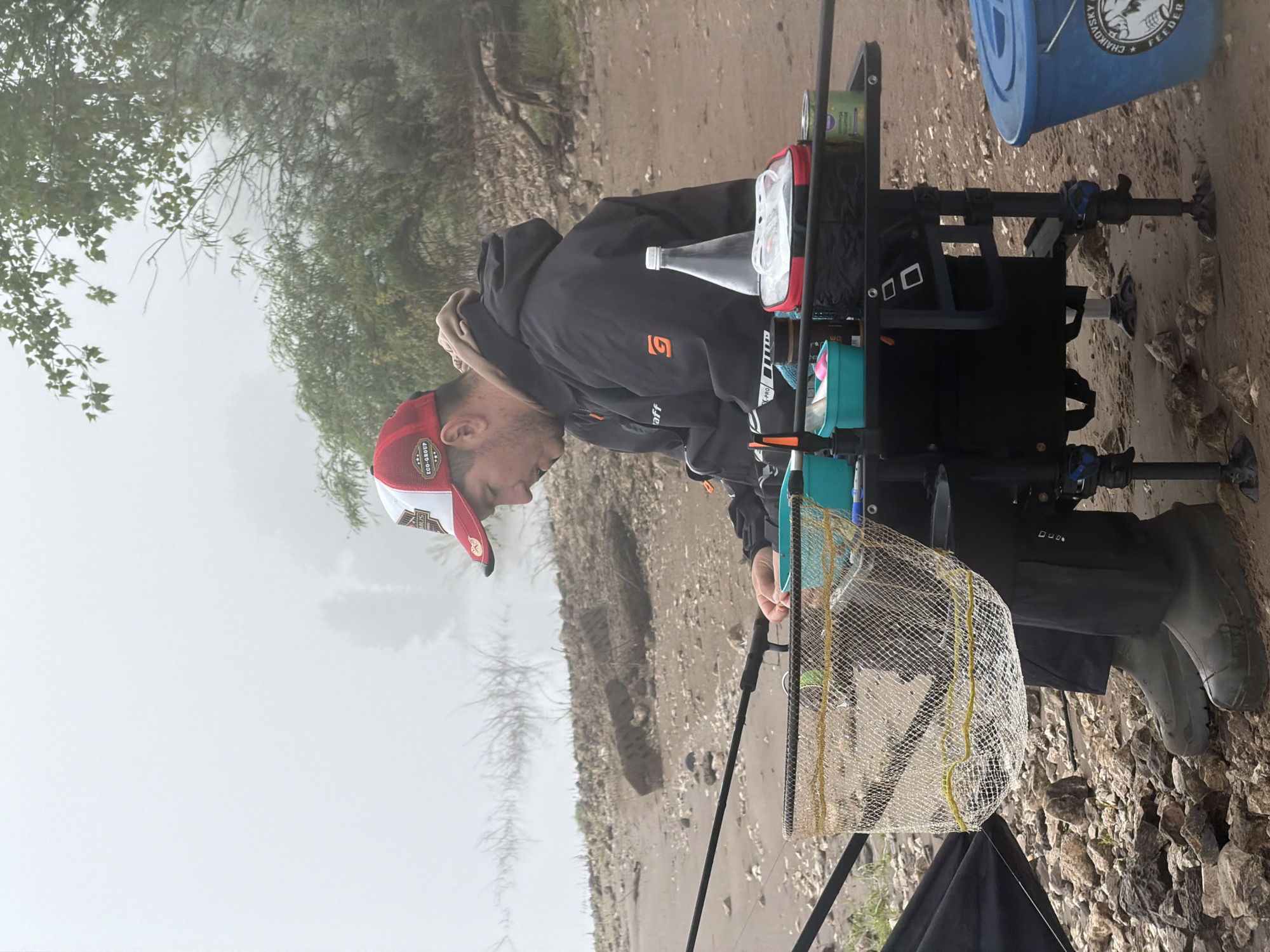 This vertical shot depicts a man engaged in fishing on a misty riverbank or lakeside, likely in Noviy, Russia. The scene conveys a quiet, focused atmosphere.
The central figure is a man, shown in profile, looking downwards with a concentrated expression. He has a beard and is dressed in a black hooded jacket featuring an orange "G" logo and the word "Gaff". Underneath, he wears a beige hoodie. A red and white cap, emblazoned with "ECO-GROUP", covers his head, and he sports dark rubber boots.
He is positioned next to a sophisticated fishing seat box or platform, which stands on multiple adjustable legs. This setup holds various fishing essentials: a clear plastic bottle with a white cap, a red and black soft case, a green container, and other smaller items. A light-colored mesh keepnet with yellow ties is attached to the platform, ready to secure any catches. Fishing rods are visible, extending into the frame, indicating active fishing.
The setting is an outdoor, natural environment characterized by a rugged bank made of dirt, sand, and scattered rocks. The background is diffused by a heavy mist or fog, through which sparse trees and shrubs are faintly visible. In the upper right corner, a blue bucket with white text reading "CHAIKOVSKY" and "FEEDER" is seen, alongside a logo featuring what appears to be a fish or sea creature. The misty conditions suggest an overcast day, possibly early morning or late afternoon.