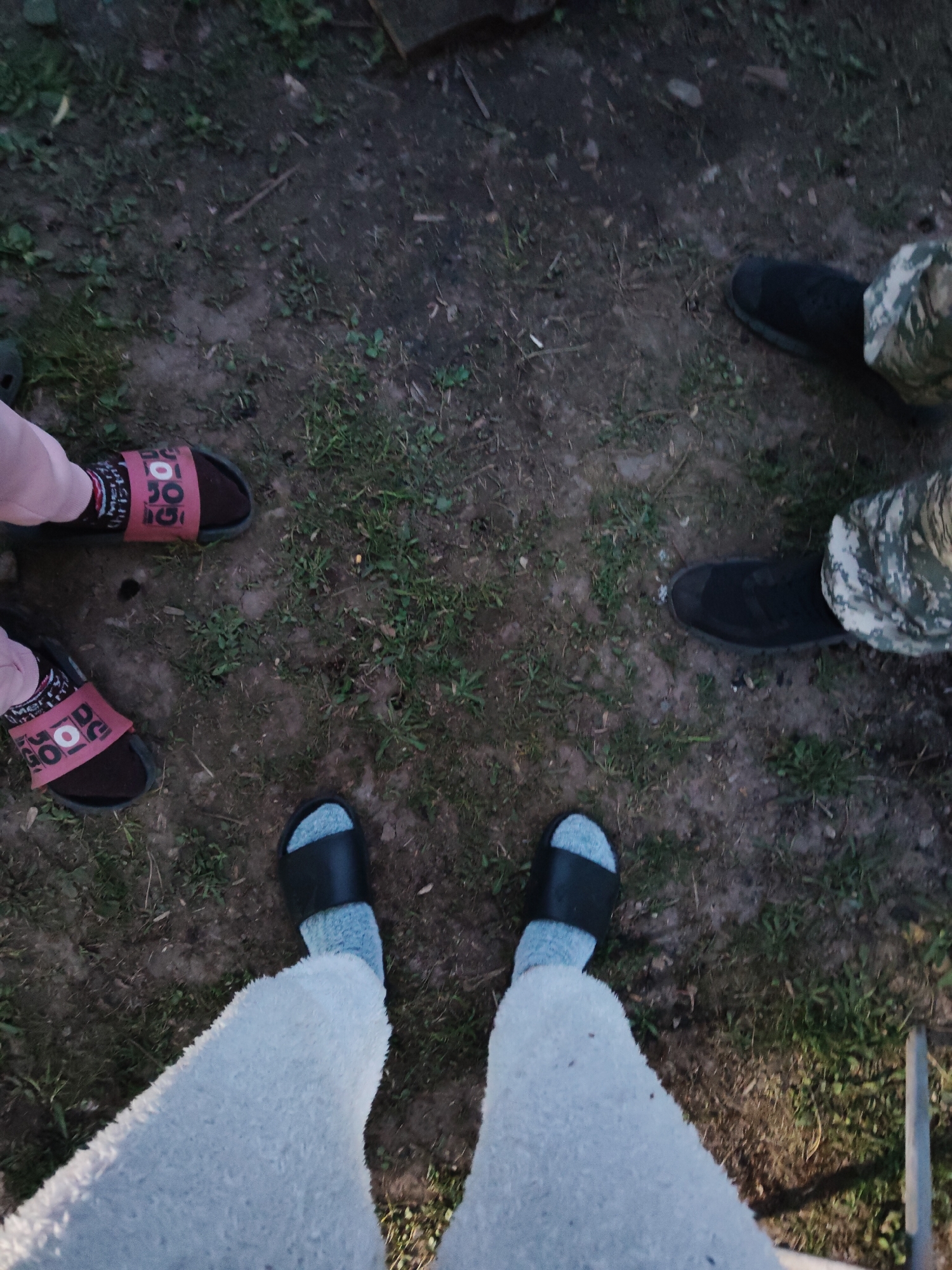 The image depicts a ground-level view of four pairs of feet standing on a dirt and grass surface. The feet are arranged in a rough circle. The individual on the left is wearing pink socks and dark-colored sandals with a possible logo or text on the strap. The person on the right is wearing dark-colored boots and camouflage pants. The person at the bottom of the image is wearing light grey socks and black slide sandals, and is also wearing what seems to be a light gray pair of fluffy pants. The ground is primarily dirt with patches of green grass. Part of a wooden structure is visible in the upper center of the image, and part of a metal structure is visible on the lower right. The overall lighting suggests an outdoor setting in twilight or dim light. Location is unknown.