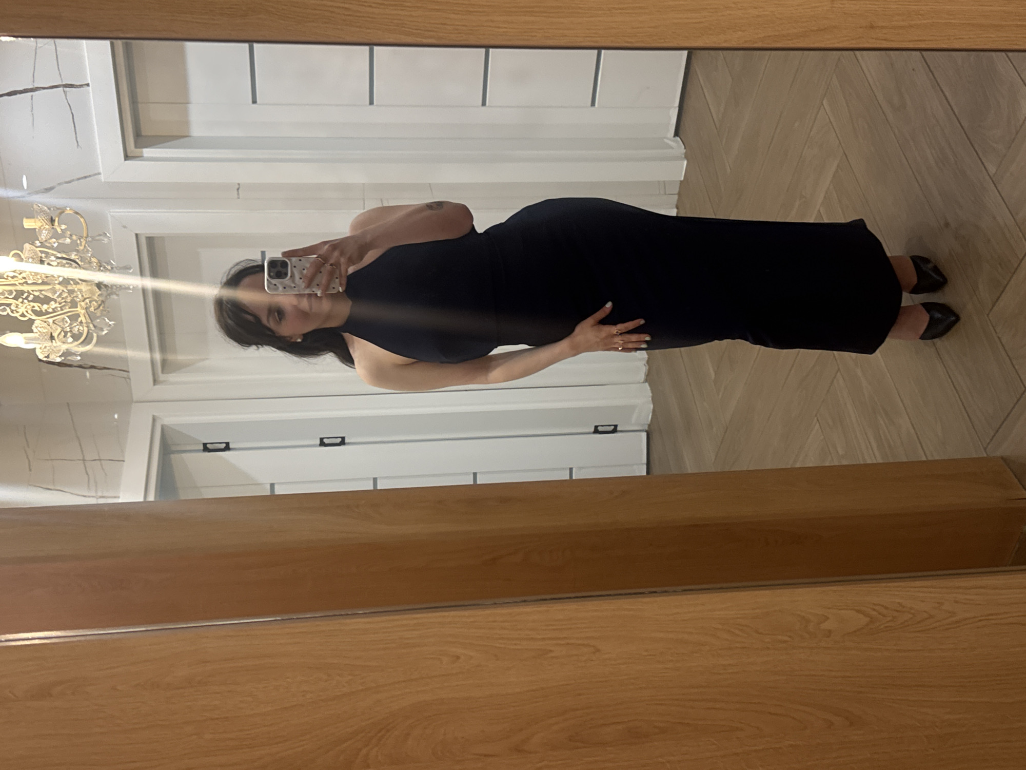 An indoor mirror selfie captures a woman posing in a well-lit room. She stands sideways to a large mirror with a thick, light-brown wooden frame, holding a white smartphone with a patterned case to take the picture.
The woman has dark hair with bangs and is wearing a form-fitting black sleeveless maxi dress and dark low-heeled shoes. Her left arm is raised to hold the phone, while her right hand rests gently on her stomach. A small, dark tattoo is visible on her upper right arm. She wears rings on her fingers, and her fingernails are painted a light color. Her facial expression is a subtle, pleasant smile.
The reflection in the mirror reveals a bright interior space. The floor is covered with light wood laid in a distinct herringbone pattern. White walls, some appearing to have subtle paneling or tile-like features, are visible behind her. An ornate, gold-colored chandelier with multiple light sources is prominently reflected on the left side of the image, contributing to the room's illumination. The scene suggests a modern and elegant setting, possibly a hallway or an entryway in a home or hotel in Blagoevgrad, Bulgaria. No legible text is visible in the image.