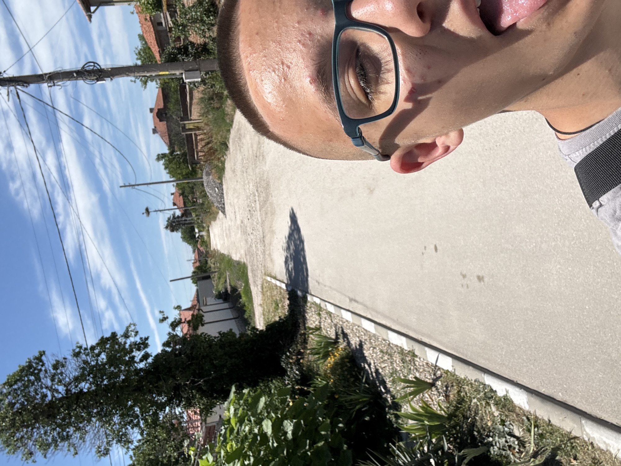 A selfie taken in Kostievo, Bulgaria, shows a person wearing blue glasses, with their tongue sticking out. The background includes a street, vegetation, utility poles, and buildings under a blue, partly cloudy sky.