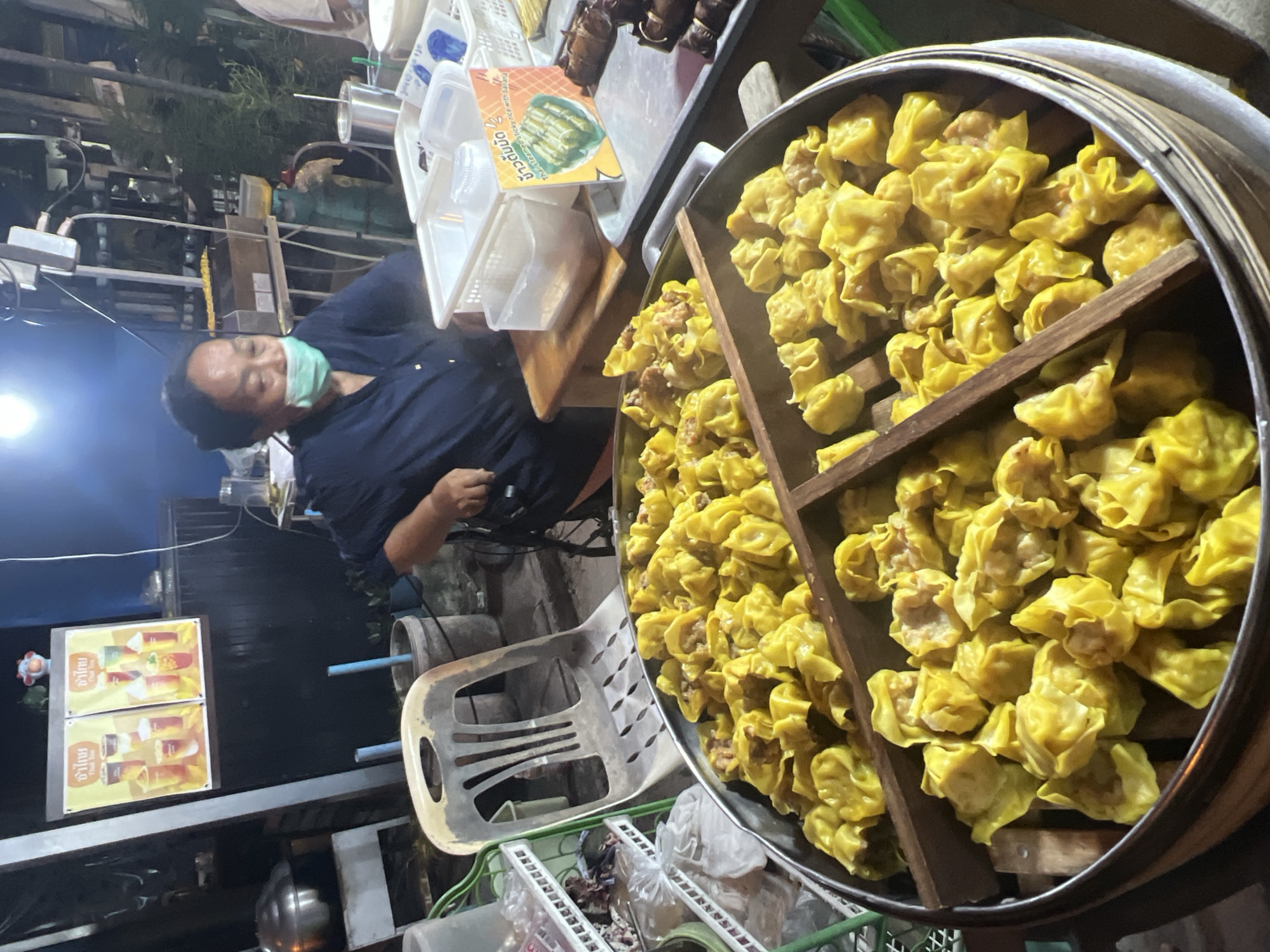 A woman wearing a face mask is serving dim sum at a street food stall at night in Ko Pha-ngan District, Thailand. She is seated behind a large steamer basket filled with yellow dim sum dumplings. In the background, there are posters advertising drinks, a chair, a gas cylinder, and other food items. The stall appears to be part of an outdoor night market or a similar food vendor setting.