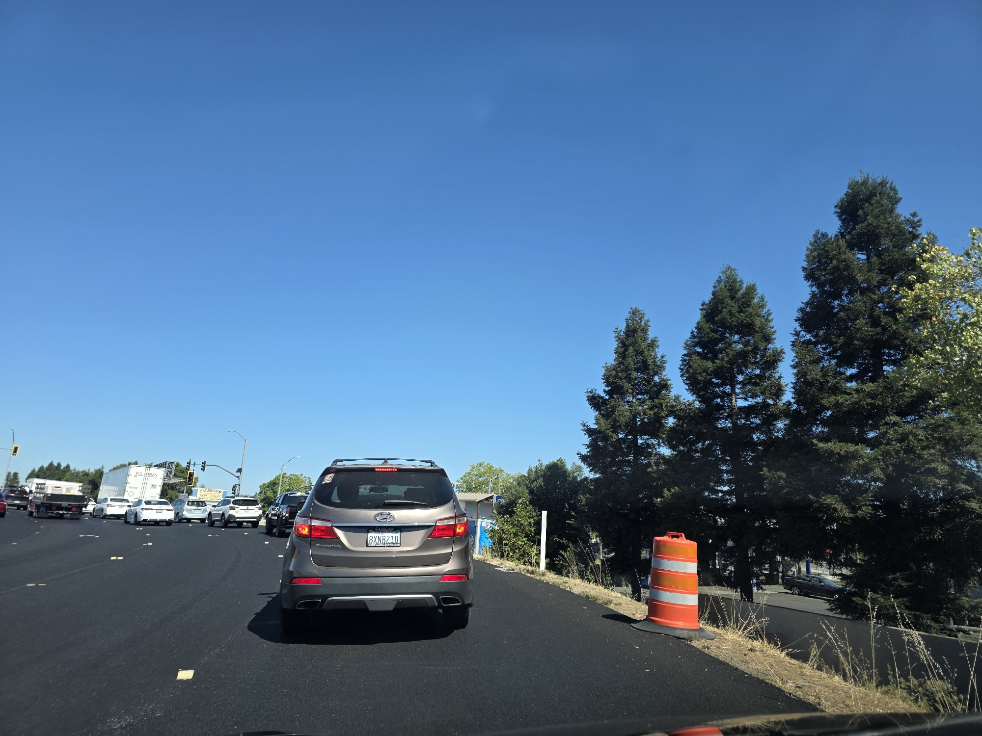 The image captures a view from the driver's seat of a car on a sunny day in West Sacramento, USA. The scene is a multi-lane road with moderate traffic. In the foreground, the rear of a brown SUV is visible, its license plate reading "8XNR210". The road surface is dark asphalt, with yellow rectangular markers.
Several other vehicles are on the road, including white cars, a white truck with a trailer, and a black SUV. In the distance, traffic lights and signs are visible. To the right of the road, there is a construction zone with an orange and white traffic barrel. Beyond that, a line of tall evergreen trees stands against a clear blue sky. The sun is bright, indicating it is likely daytime, and the clear sky suggests fair weather. The overall impression is one of a typical commute or travel on a highway. There is no discernible emotion conveyed by the image.
Visible text includes the license plate "8XNR210" on the SUV, and text on signs in the distance, though the text on the signs is illegible. A white trailer in the distance has text on its side, and a yellow sign also has text that is too blurry to read.