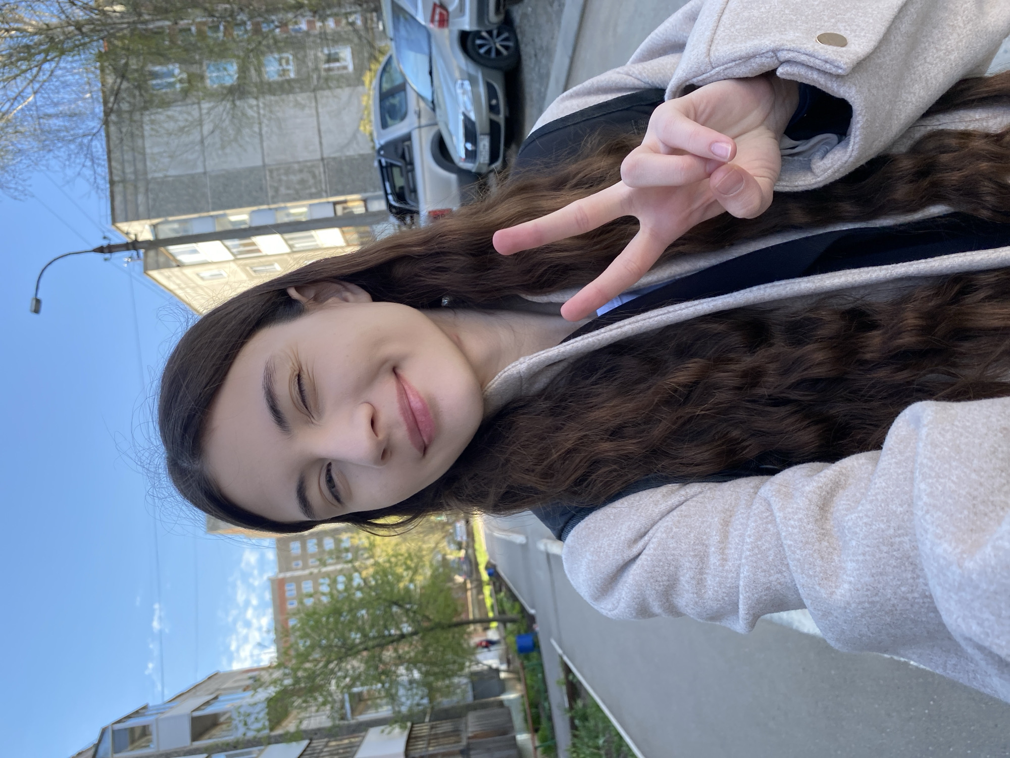 A young woman is taking a selfie outdoors. She is wearing a light-colored coat with a black and blue top beneath. She is smiling and winking at the camera, holding up a peace sign with her right hand. Her hair is long and wavy. In the background, there are apartment buildings and cars. The sky is blue. The location appears to be Votkinsk, Russia.
