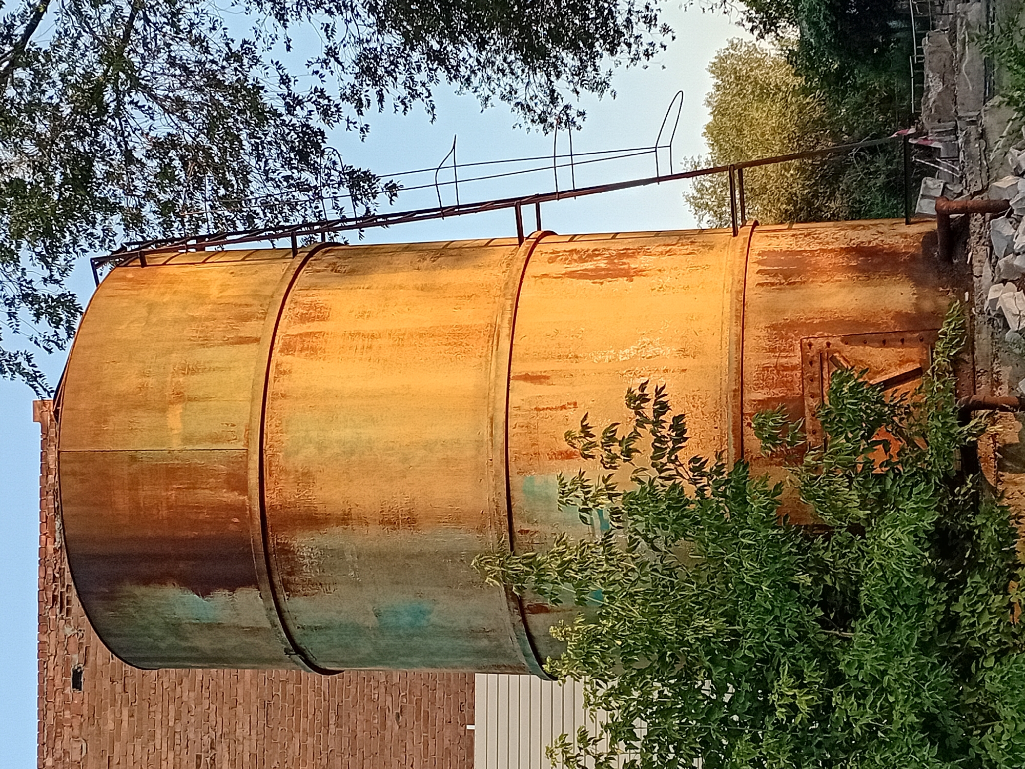 The image depicts a tall, weathered cylindrical metal tank, likely a water tower or silo, located in the city of Semey, Kazakhstan. The tank exhibits significant rust and patches of faded green paint, indicating age and exposure to the elements. A metal ladder with safety cages extends up the side of the tank. In the foreground, lush green foliage partially obscures the base of the tank. Behind the tank, a brick building with a light-colored section at its base is visible, suggesting an industrial or older urban setting. The sky is clear and blue, and dappled sunlight illuminates the tank, casting warm tones on its surface. The surrounding environment includes trees and some debris at the base, hinting at a semi-developed or possibly neglected area. There are no people visible in the image.