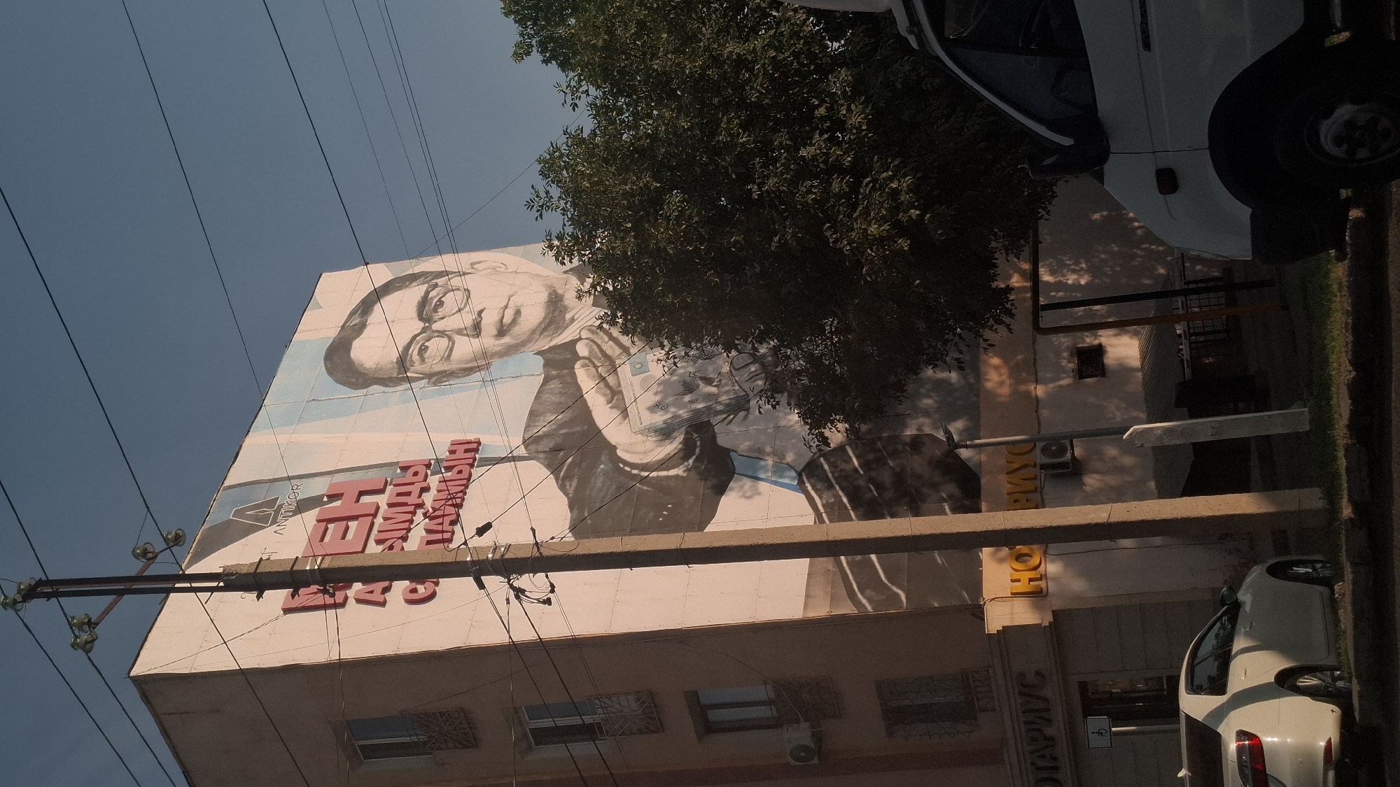 The image shows a large mural on the side of a building in Shymkent, Kazakhstan. The mural depicts a man in a suit wearing glasses, holding a wad of money. To the left of the mural, large red Cyrillic letters spell out a message. In the foreground, a white car is parked on the side of the street, and a portion of a white van is visible on the right side of the image. A utility pole with wires runs vertically through the scene, obscuring part of the mural. Trees are present on the right side of the image, casting shadows on the building. Signs for "НОТАРИУС" (NOTARY) are visible on the lower part of the building. The sky is a clear blue.