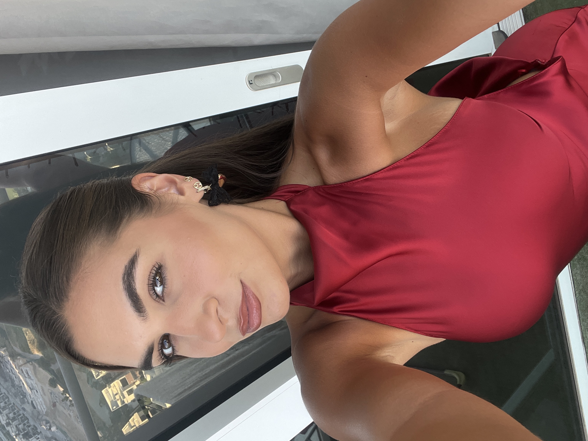 A woman with dark hair is posing in what appears to be a selfie. She is wearing a reddish-brown dress. She is facing the camera with her head tilted, and she has a black earring in her left ear. The background shows a view of a city skyline through a window. Based on the architectural style and the presence of skyscrapers, the scene may be set in Dubai, United Arab Emirates.