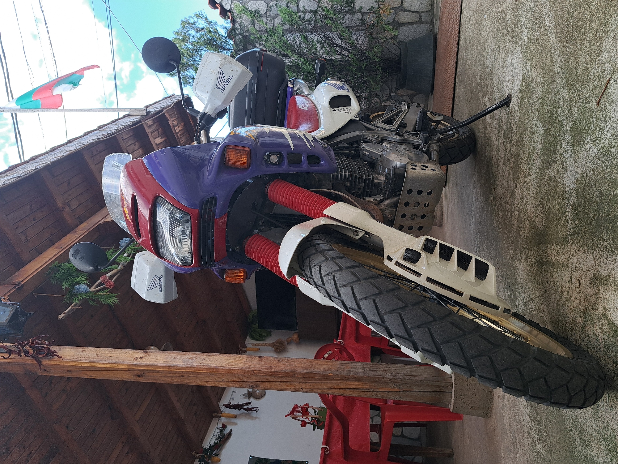 A motorcycle is parked on a paved surface, possibly concrete. The motorcycle has a distinctive red, white, and purple color scheme, and appears to be an older model with a large front tire. The motorcycle is positioned near a structure with a wooden beam and tiled roof. In the background, there is a stone wall and some greenery. Above the roof, a flag is visible with white, green and red stripes. The motorcycle is a Honda.