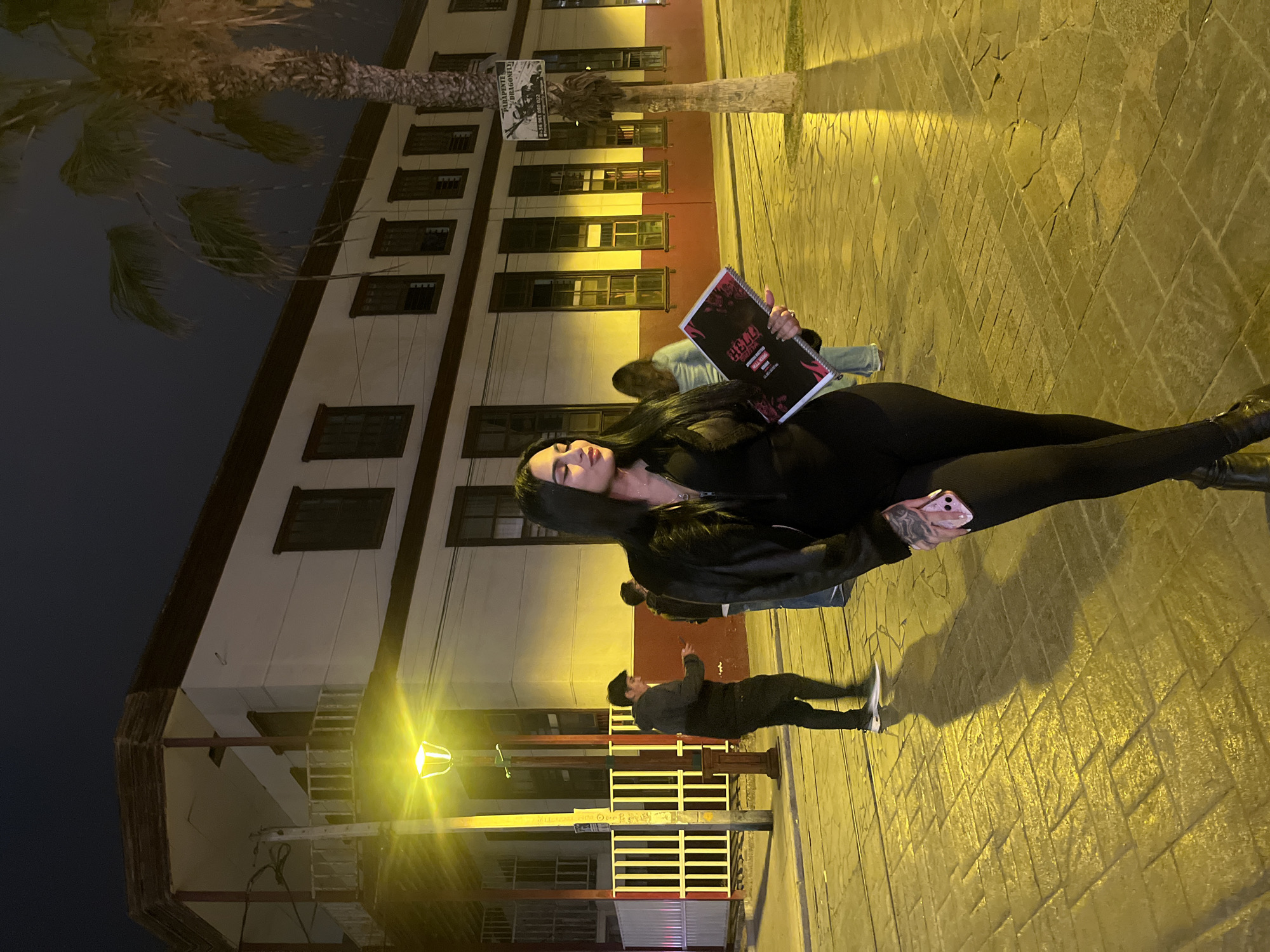 A woman is standing on a cobblestone street at night, holding a brochure or menu. She is wearing a black outfit and has long dark hair. In the background, there is a building with multiple windows and a palm tree. The street is illuminated by a yellow streetlight. Other people are visible in the background, walking on the street. The scene appears to be in the city of Iquique, Chile, based on the architecture and vegetation.