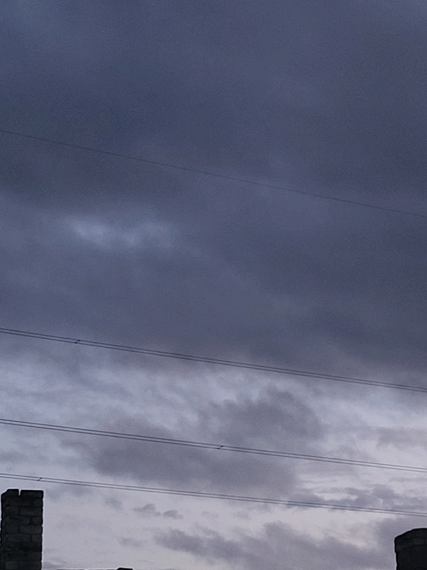 The image captures a grey, overcast sky with horizontal power lines stretching across the frame. In the foreground, the tops of two brick structures are visible, one on the left and another on the right, partially silhouetted against the muted sky. The structure on the left appears to be a chimney or a section of a building, made of dark bricks. The structure on the right is less defined but also appears to be a brick construction. The overall scene suggests an outdoor environment, likely an urban or industrial setting in Jelgava, Latvia, under a gloomy sky. There are no people or animals visible in the image. The composition is dominated by the sky and the power lines, with the brick structures providing a grounding element at the bottom of the frame.