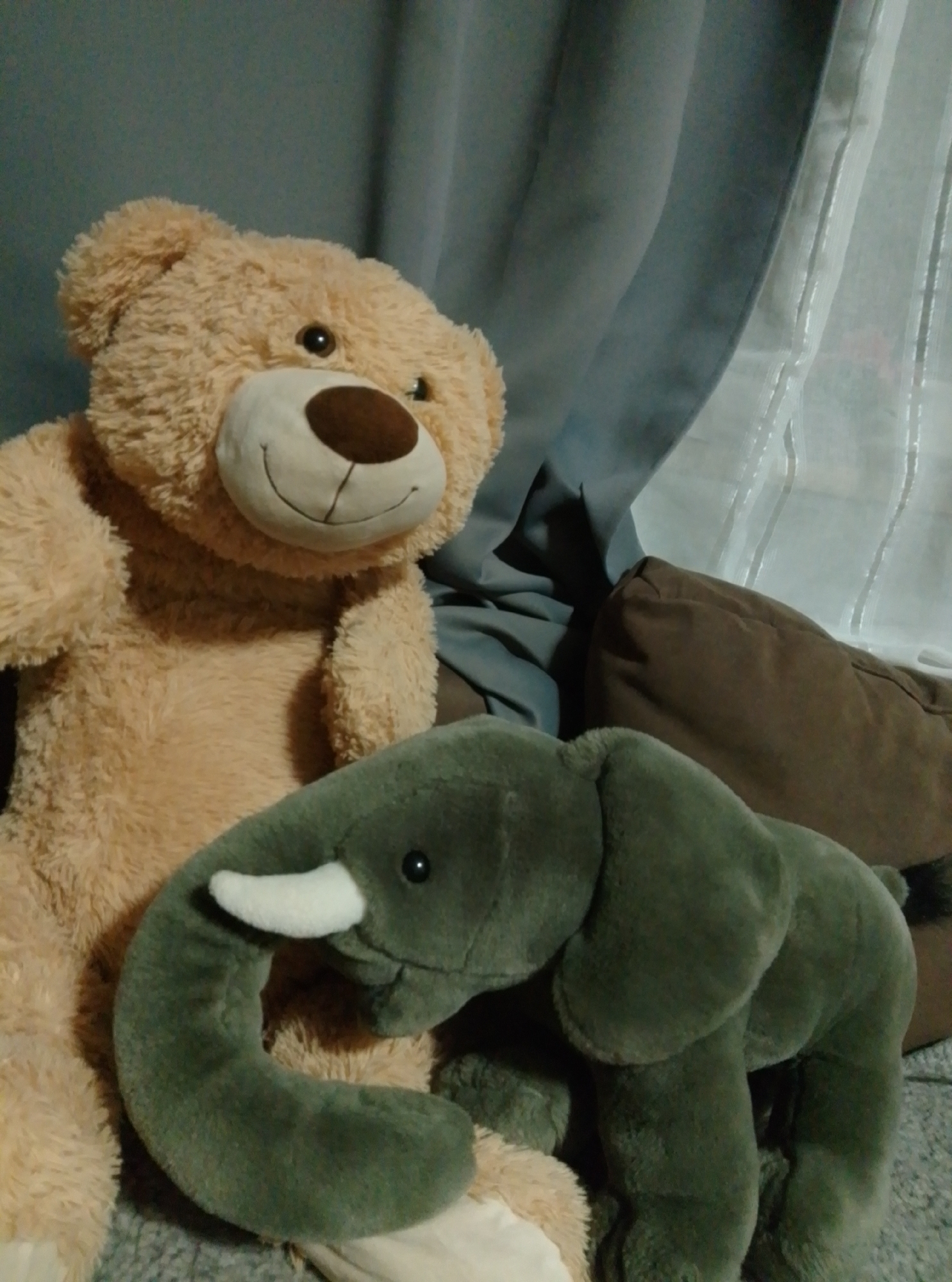 The image shows two stuffed toys: a large, light brown teddy bear and a smaller, dark green elephant. The teddy bear is positioned on the left, with its head and upper body visible. It has dark eyes, a brown nose, and a stitched smile. The elephant is in the foreground on the right, with its trunk curved upwards and one white tusk visible. Its eyes are dark and beady. They are both placed on a textured surface that appears to be a blanket or rug, possibly on a bed or sofa. In the background, there are draped curtains, one a solid blue-grey and the other a sheer white with vertical stripes. The lighting is dim, suggesting an indoor setting, possibly at night or in a room with closed blinds. There is no visible indication of the city of Wedel, Germany.