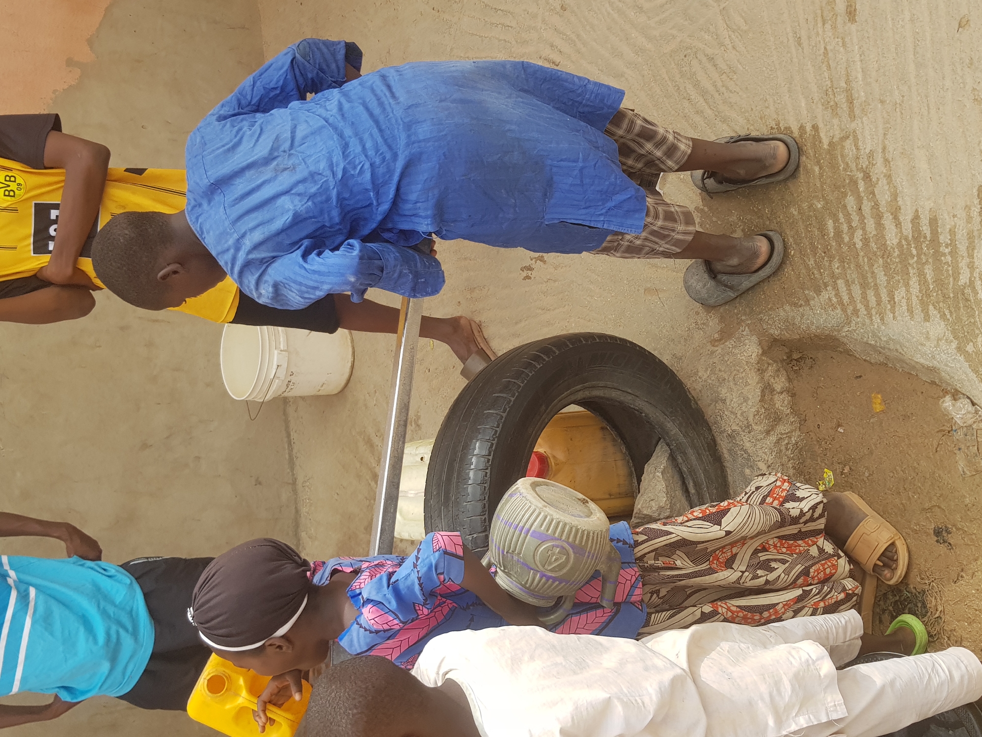 The image depicts a group of people outdoors in what appears to be a semi-urban area, potentially in Dutsinma B, Nigeria. Several individuals are gathered around a tire and various containers, possibly involved in a water-related activity. One person, wearing a blue garment and plaid shorts, leans over a metal pole positioned near a white bucket. Another person, wearing a blue and pink patterned shirt, rests a plastic pitcher on a white garment. Other people are visible wearing different colored shirts. The environment consists of a concrete surface and what looks like a wall in the background.