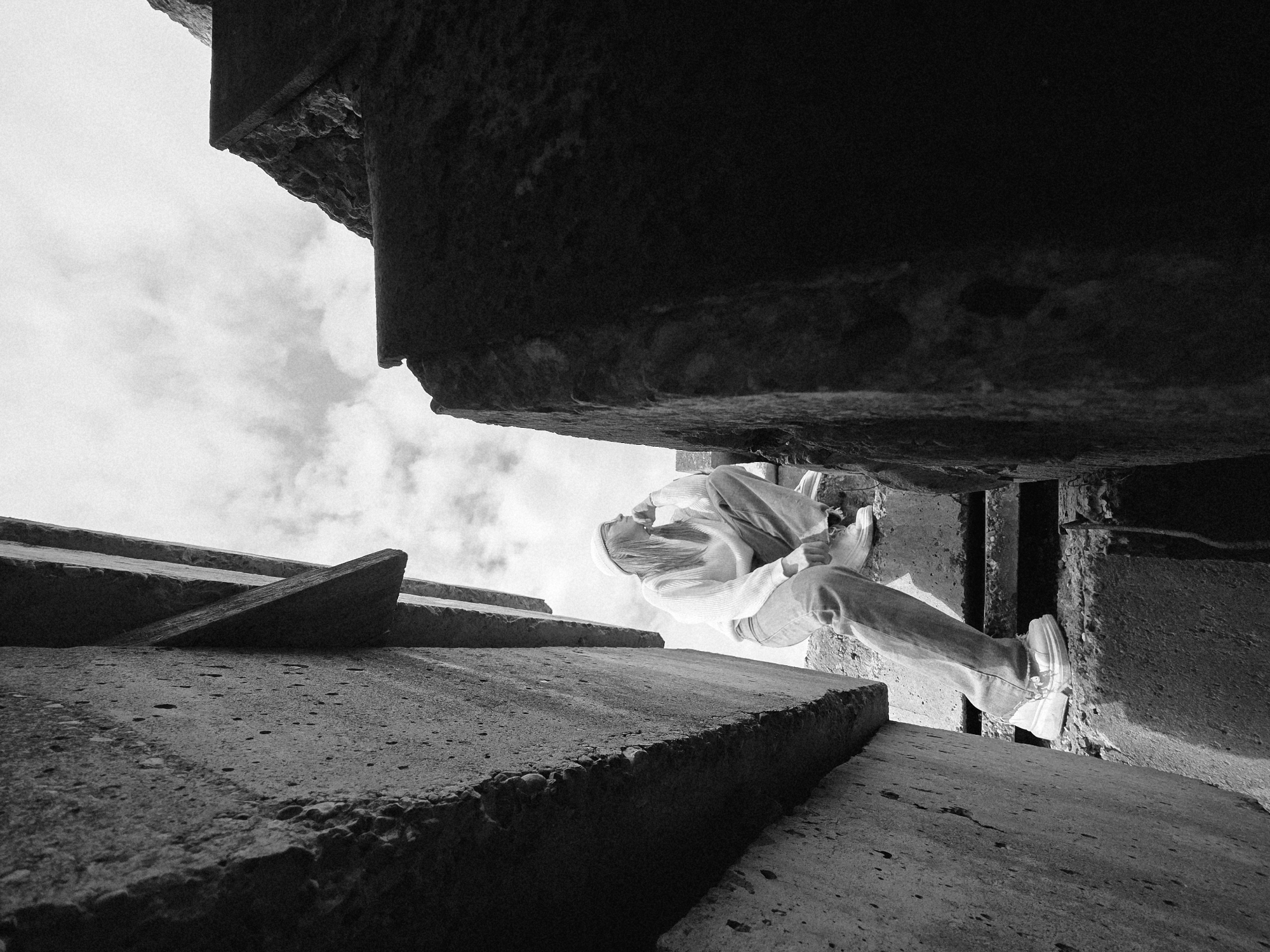 A low-angle, black and white photograph depicts a person sitting within a concrete structure, possibly stairs or retaining walls, against a cloudy sky. The person, seen from the waist down, is wearing light-colored jeans and white sneakers. They appear to be perched on the edge of one of the concrete elements. The scene is composed of geometric forms created by the concrete, emphasizing texture and perspective. The location is described as "Unknown, Unknown."