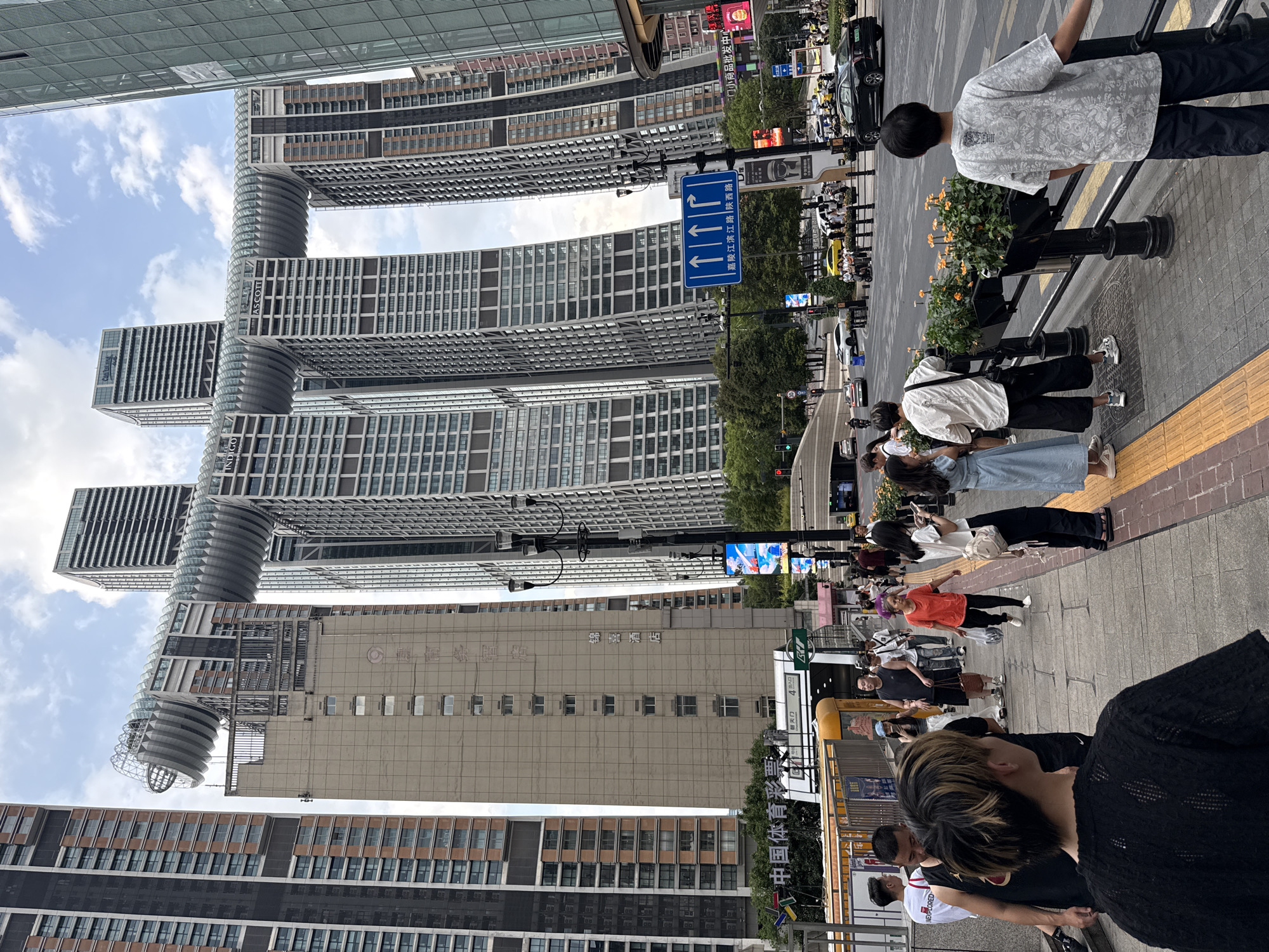 In the Yuzhong District of China, the scene captures a bustling urban environment dominated by modern skyscrapers. The most prominent structures are a group of interconnected buildings featuring a unique, wave-like, transparent walkway spanning across their upper levels, appearing as a colossal bridge. The architecture is characterized by sleek lines and glass facades. In the foreground, a sidewalk is populated by a diverse group of pedestrians. Some are walking, others are standing and conversing, and one individual in the immediate foreground is facing away from the camera, looking towards the street. The sidewalk is bordered by planters filled with orange flowers. A busy street with multiple lanes of traffic runs alongside the sidewalk, with various vehicles, including cars, motorcycles, and a yellow bus, visible. Traffic lights and street signs are present. The sky above is partly cloudy, indicating daytime. The overall impression is one of a vibrant and dynamic city district with striking contemporary architecture and everyday urban activity.