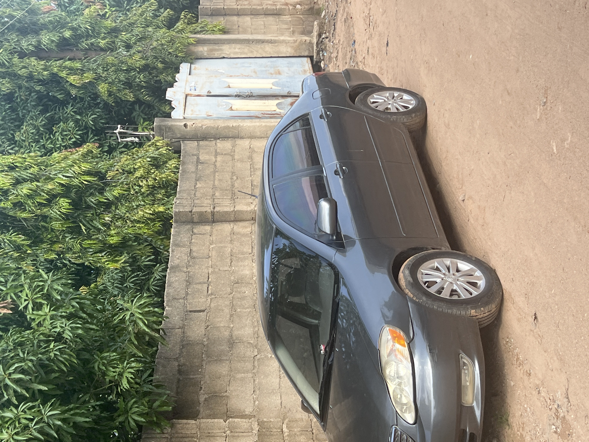 In Jalingo, Nigeria, a dark gray sedan is parked on a dirt road next to a rough, cinder block wall. Lush green trees with dense foliage line the top of the wall, creating a natural backdrop. To the right of the wall, a large, weathered, light blue metal gate stands partially open, revealing more of the vegetation behind it. The car's front passenger side is visible, showing its headlight, bumper, and a silver alloy wheel. The road surface is uneven and dusty, with scattered debris. The lighting suggests it is daytime.