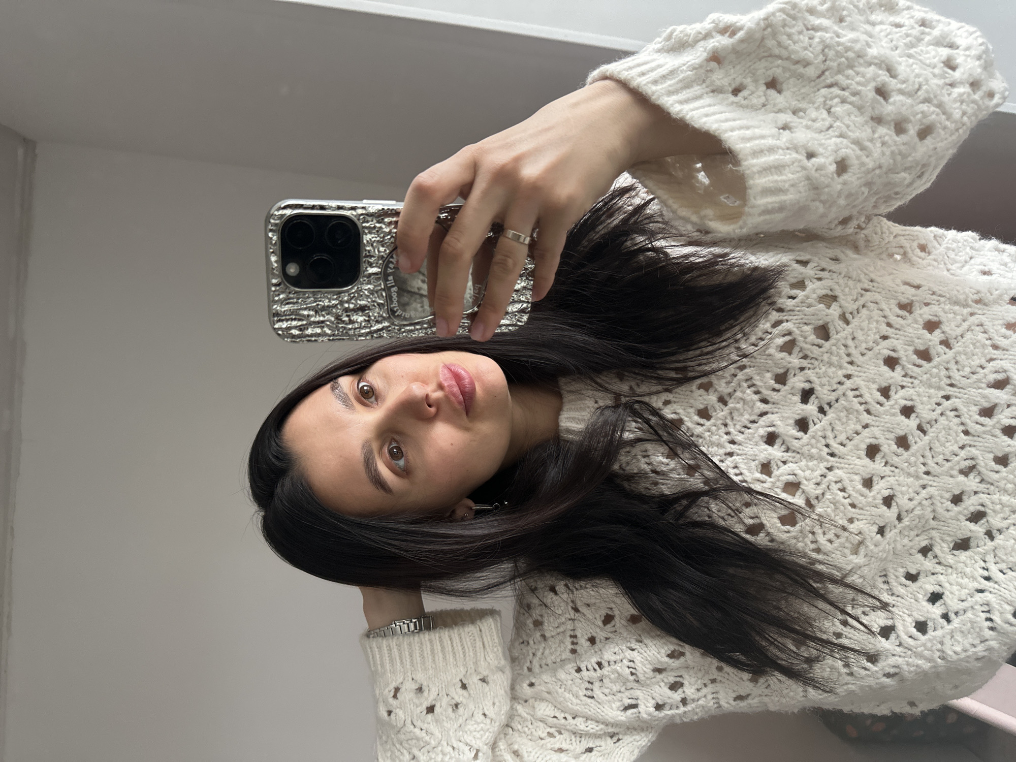 A woman with long, dark hair is taking a selfie in a room with light-colored walls. She is wearing a chunky knit, cream-colored sweater and a silver watch. She is holding a silver smartphone with multiple camera lenses. A ring is visible on one of her fingers. Her location in Chaikovsky, Russia is not discernible from the image.