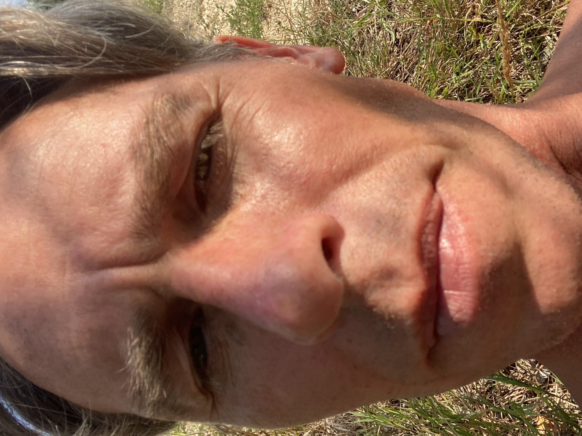 A close-up, low-angle, selfie-style shot features a fair-skinned man with graying hair and faint wrinkles on his forehead and around his eyes. His left eye is partially obscured by shadow. The man's skin appears flushed, possibly from sun exposure. He is outdoors, with dry grass and some taller stalks visible in the background. The lighting suggests it is daytime, with sunlight casting shadows on his face. The location is identified as Куйбышевское сельское поселение, Ukraine.