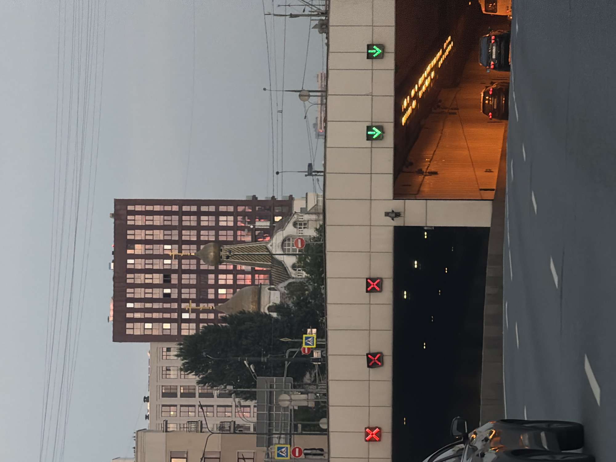 The image shows an urban scene in Moscow, Russia, at dusk. In the foreground, a road with white lane markings is visible, with the rear of a dark-colored car on the left. Above the road, a large concrete structure with a tunnel entrance spans the width of the frame. Red illuminated "X" signs are displayed above the tunnel entrance, indicating closure or prohibition. To the right of the red "X" signs, green downward-pointing arrows are visible, presumably indicating open lanes or directions.
Behind the concrete structure, the cityscape features several buildings. A prominent, tall, reddish-brown building with a grid of illuminated windows dominates the middle ground. To its left, a cluster of lighter-colored apartment buildings with similar window patterns can be seen. Between these buildings, a traditional Orthodox church with golden domes and crosses stands out. Trees are also present in this area. The sky is overcast and a pale gray, with power lines crisscrossing the upper portion of the image. The overall impression is one of a busy urban environment at the transition from day to night, with traffic infrastructure and religious architecture coexisting.
