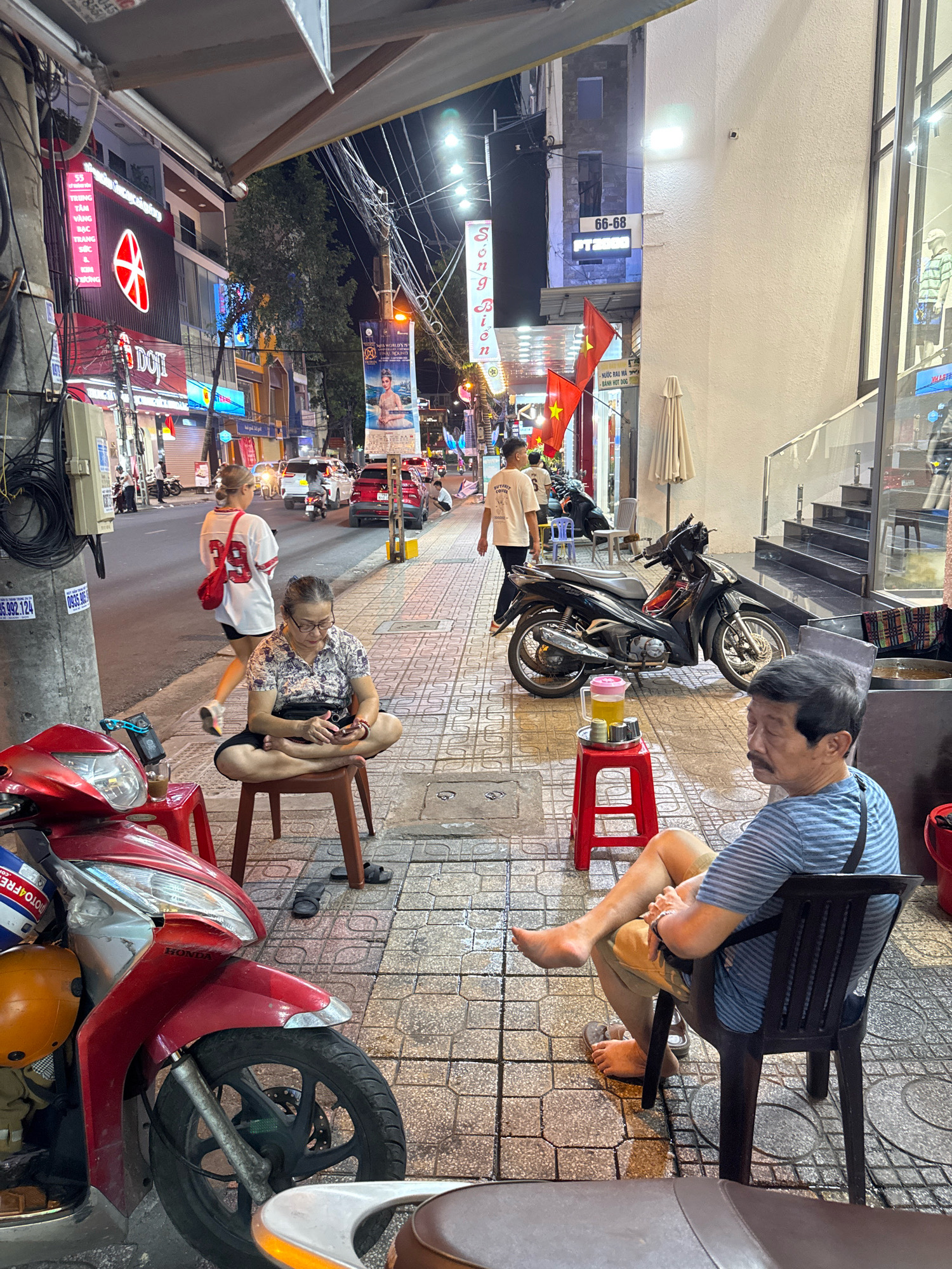 This image depicts a street scene at night in Nha Trang, Vietnam. A woman is seated on a stool on the sidewalk, facing a smartphone. She is wearing glasses and a patterned shirt, with her legs crossed in a meditative pose. To her right, a black motorcycle is parked. Further down the sidewalk, a man is seated in a plastic chair, his legs crossed and his gaze directed away from the camera. He is wearing a striped t-shirt and shorts. A tall glass of yellow liquid with a pink lid sits on a red stool near the motorcycle. Red scooters are parked in the foreground. The street is paved with patterned tiles, and buildings with signs and lights line the street. Several streetlights illuminate the scene.