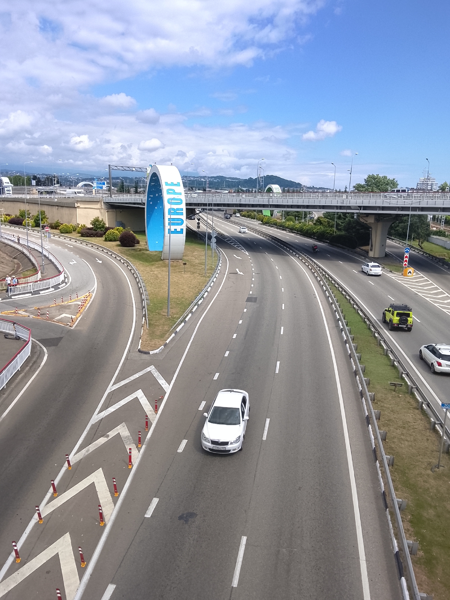 This media file depicts a view of a multi-lane highway under a bright, partly cloudy sky, suggesting daytime. The scene is set on a wide, paved roadway with clear lane markings. A prominent architectural element, a large, blue, arch-like structure with the word "EUROPE" written vertically in white letters, stands to the left of the highway.
Several cars are visible on the highway, including a white sedan in the foreground, a yellow SUV, and other vehicles further down the road. The highway is bordered by guardrails and grassy verges. In the background, an elevated pedestrian bridge crosses the highway, and beyond that, hills and buildings are visible. The overall impression is one of an open, modern transportation hub, possibly a border crossing or a significant junction. The weather appears pleasant, with the sun shining through scattered clouds. There are no discernible people actively engaged in an event, though a few individuals are seen near the "EUROPE" structure and on the side of the road. The location cues, such as the "EUROPE" sign, suggest a place of geographical significance.