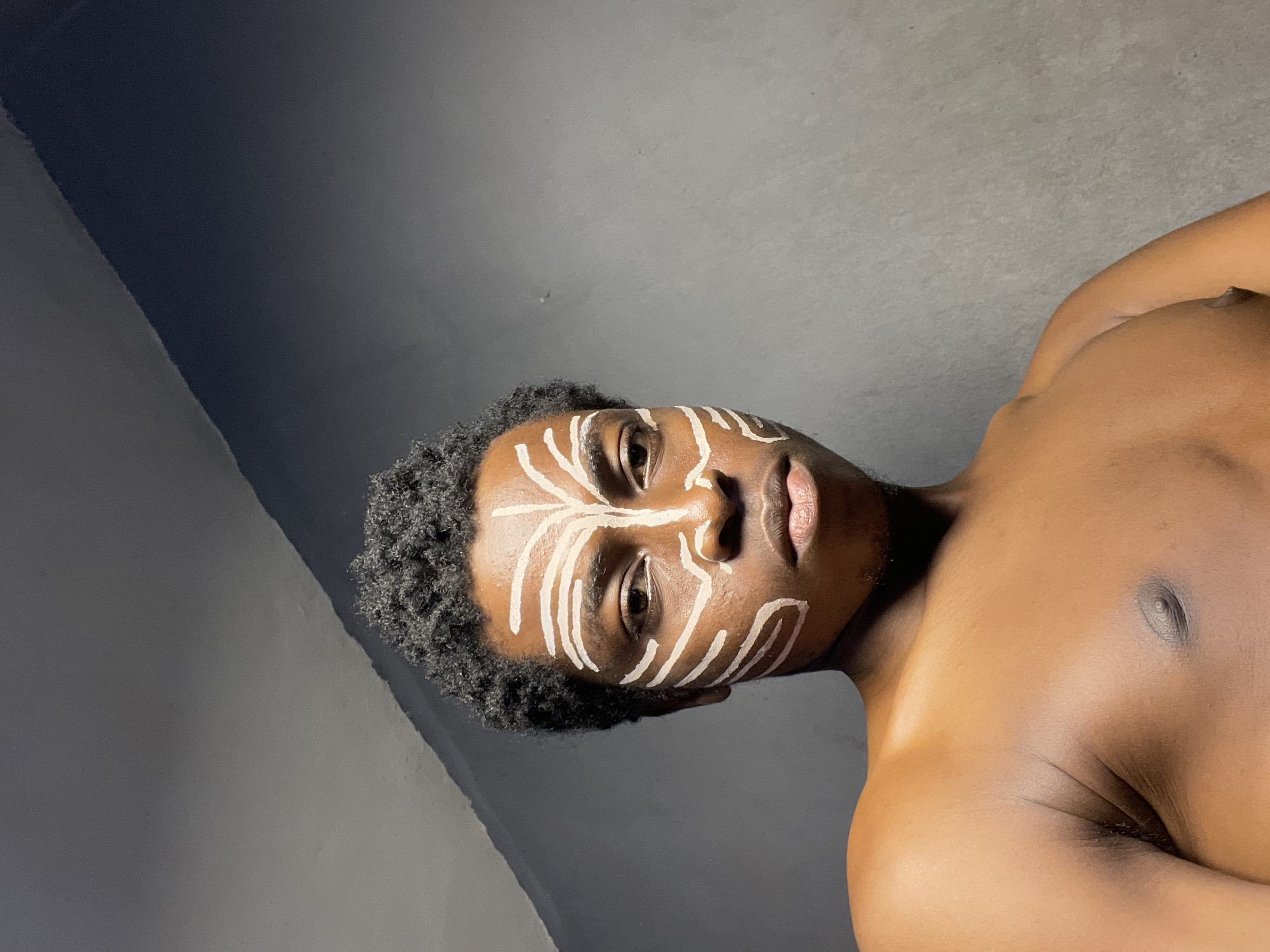 A person with dark skin and short, curly black hair is shown from the chest up, facing the camera directly. They have white face paint applied in a pattern resembling tribal markings. The markings are symmetrical, with lines extending from the center of the forehead down the bridge of the nose, around the eyes, and across the cheeks and jawline. The person's chest and shoulders are visible and appear bare. The background is a plain, textured gray wall. The image is lit from the left, casting shadows on the right side of the face and body. The person's expression is neutral. This scene appears to be an indoor setting, possibly a room in Nkpor, Nigeria, given the information.