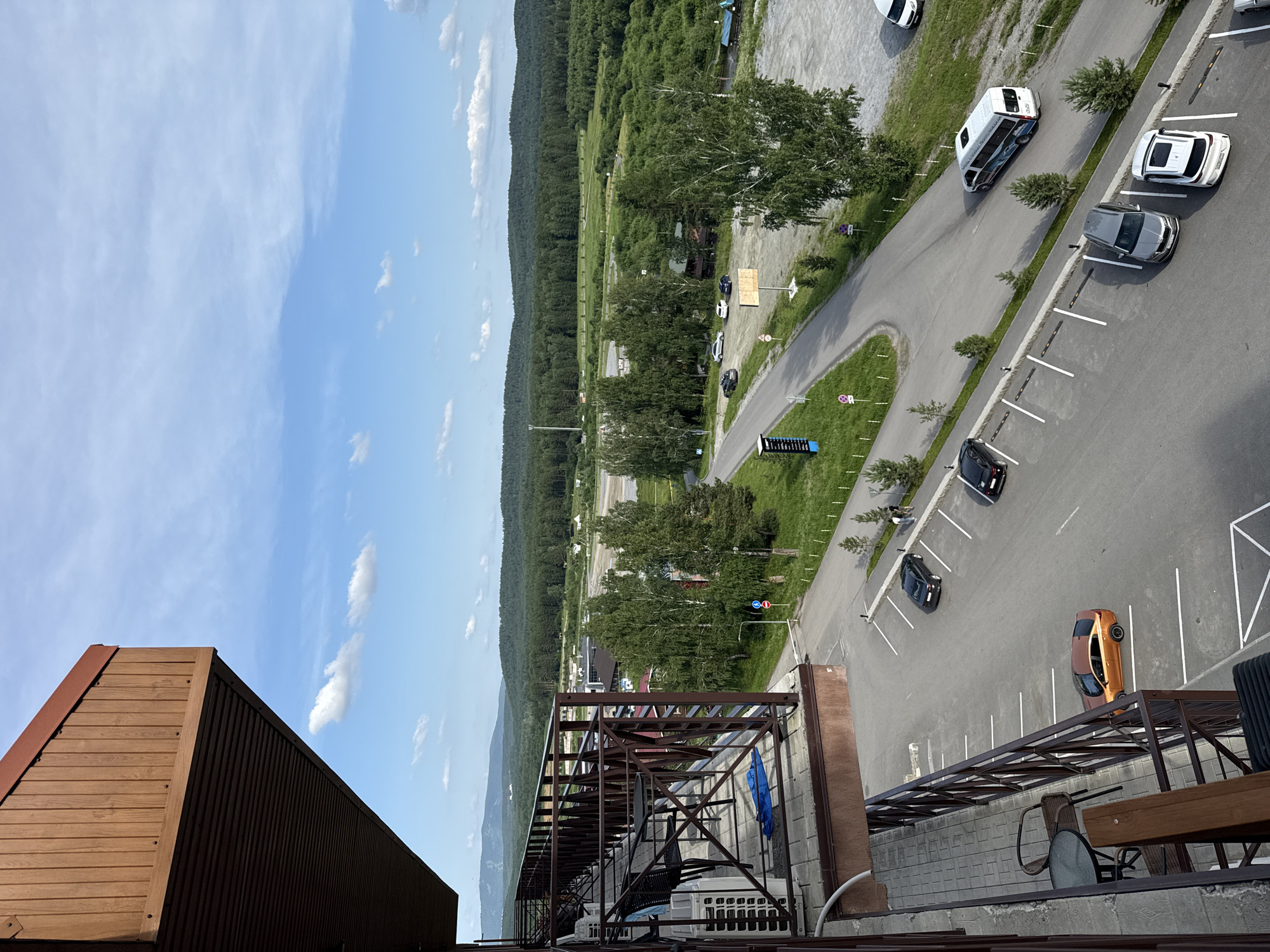 A high-angle shot captures a parking lot and surrounding landscape in Syrostan, Russia. The foreground is dominated by a balcony with a wooden railing and outdoor furniture. Below, a paved parking lot is visible with several cars parked, including a gold sedan, a dark sedan, a silver sedan, and a white SUV. A white minibus with blue graphics is driving in the parking lot. Beyond the parking lot, a paved road winds through a green area with scattered trees. In the distance, a dense forest stretches across rolling hills under a partly cloudy blue sky. The overall scene depicts a tranquil, semi-rural environment with modern infrastructure.