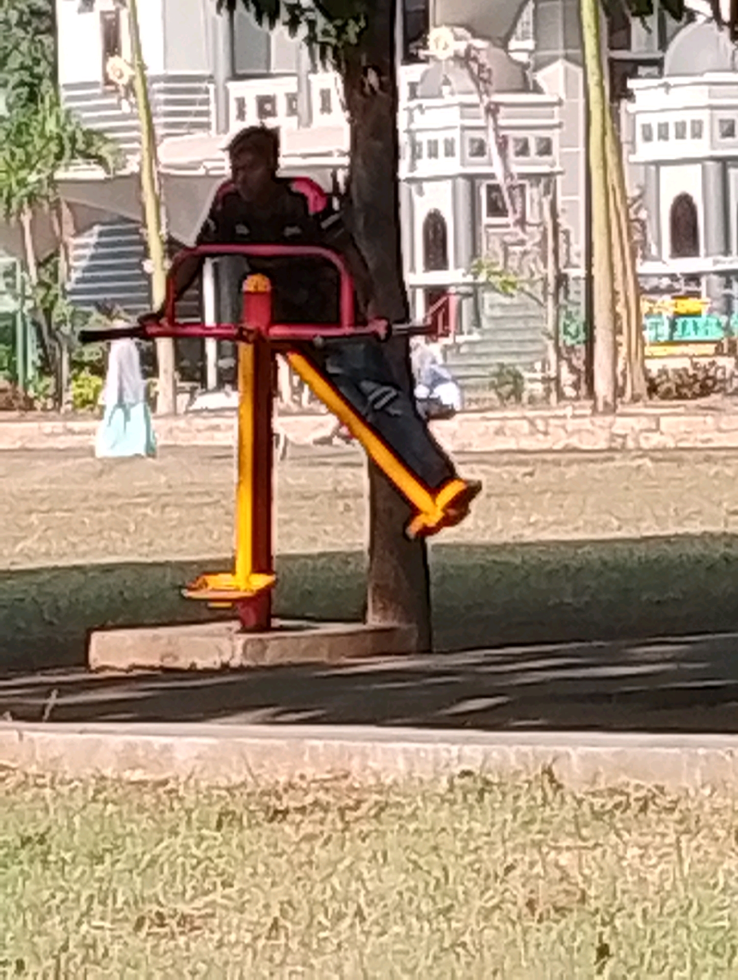 A full shot captures a man using an outdoor exercise machine in Probolinggo, Indonesia. The man, wearing a dark shirt with red and white accents, is seated on a bright red and yellow apparatus. His legs are extended forward, and he appears to be in motion. The machine has a central yellow support column. In the background, there is a grassy field, a paved walkway, and buildings with a distinct architectural style, possibly part of a recreational area or park. Trees with green foliage are also visible. The lighting suggests it is daytime.