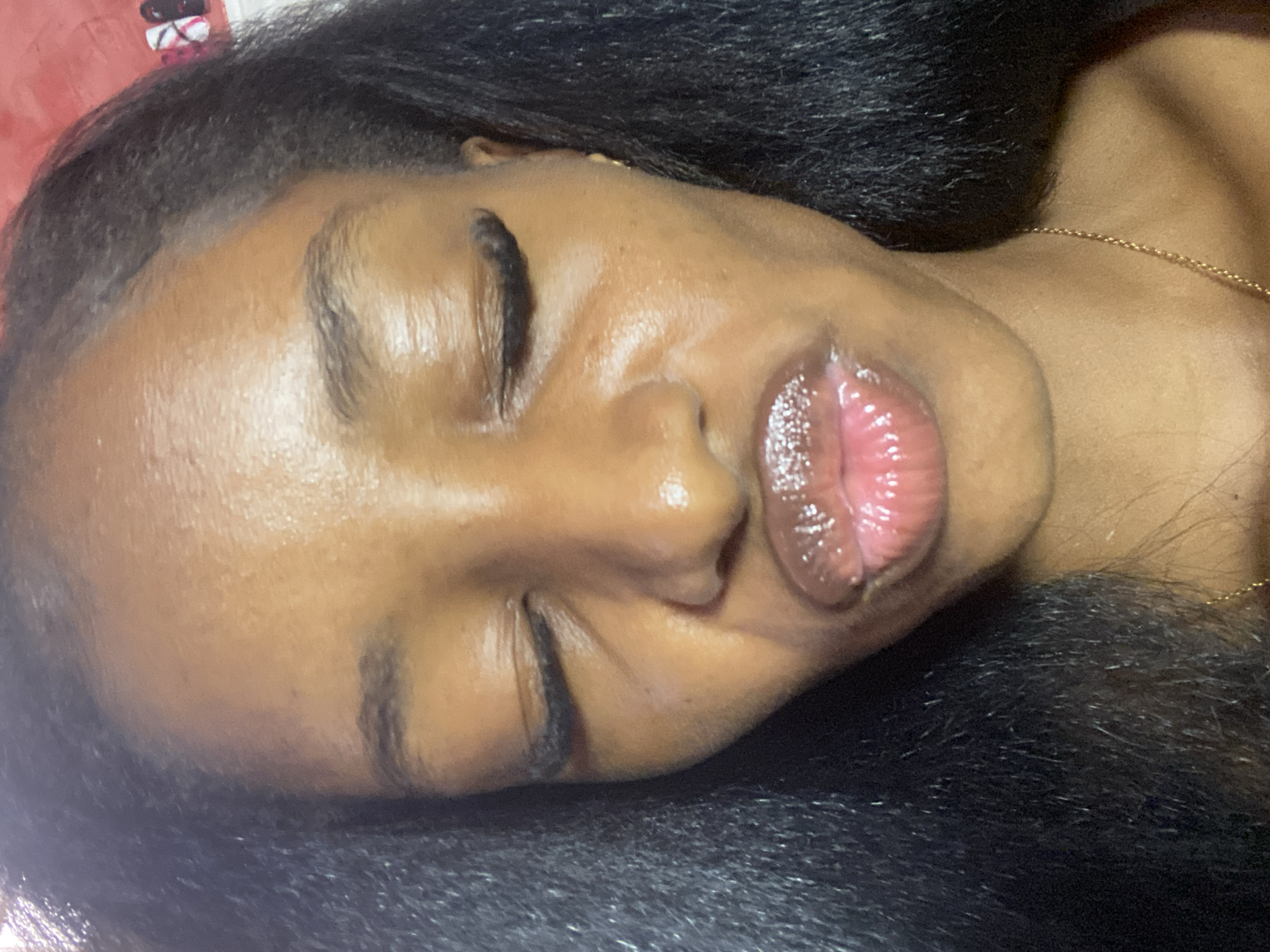 The image is a close-up, selfie-style photograph of a young Black woman's face, tilted slightly upwards, with her eyes closed and lips pursed as if blowing a kiss. She has dark, straight hair framing her face and shoulders. Her skin appears to have a dewy sheen, and she is wearing lip gloss, which catches the light. She also has noticeable eyelashes, possibly false ones, and a delicate gold necklace is visible around her neck. The background is out of focus and appears to be a room with some colored objects, possibly decorative items or clothing, on the right side. There is no discernible indication of the city of Jos, Nigeria, or any specific urban or environmental context within the image.