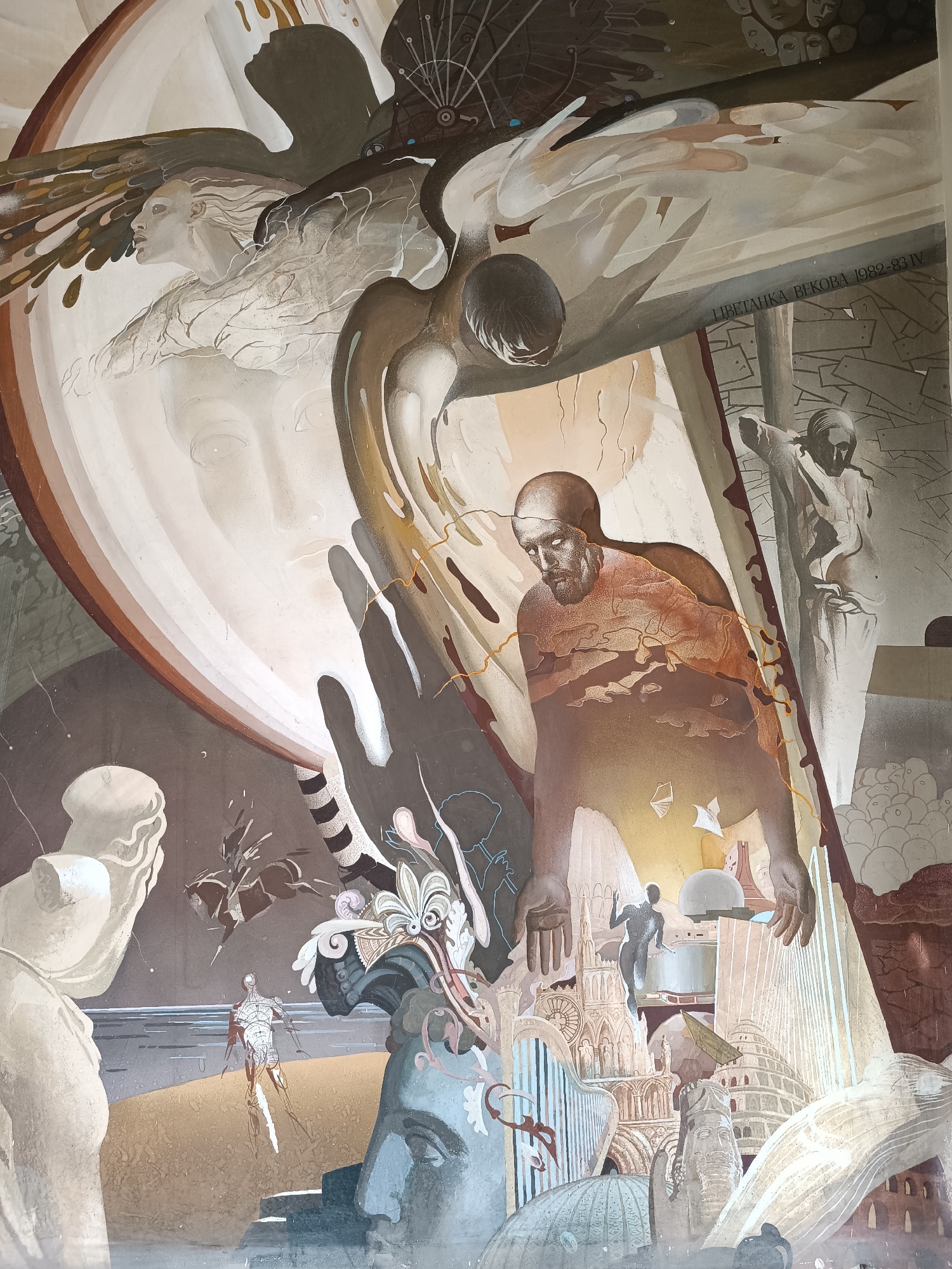 This is a photograph of a mural in Sofia, Bulgaria. The mural has a surrealist and allegorical style, featuring a complex arrangement of figures and objects.
At the top left is a large face in profile, enclosed in concentric oval shapes. Above it is an angel-like figure with wings. To the right of this, there is a dark-winged figure.
Below these is a standing man with a beard, whose torso is partly translucent, revealing a landscape within. His hands are extended downward.
The lower section of the mural includes a number of architectural structures and sculptures, partially obscured and blended together. These include a statue head, columns, the Colosseum, and other buildings. Also present are a figure walking on what appears to be a shore, a rearing horse, and a number of other indistinct figures.
A text written in the Cyrillic alphabet is visible to the right of the face.