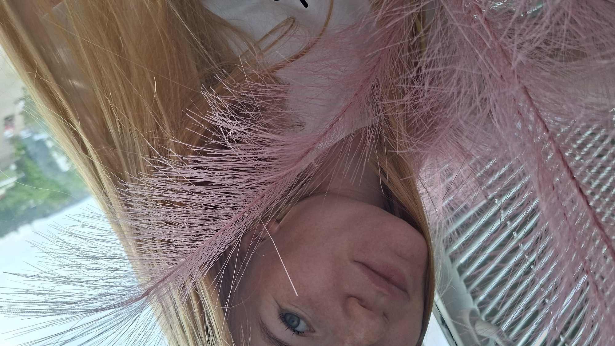 The image shows a close-up of a young woman with blonde hair and blue eyes, holding a pink feather in front of her face. She is wearing a white t-shirt. The background is out of focus, but appears to be a cityscape with buildings and trees visible. It is not possible to determine the exact location in Chaikovsky, Russia from the image alone.