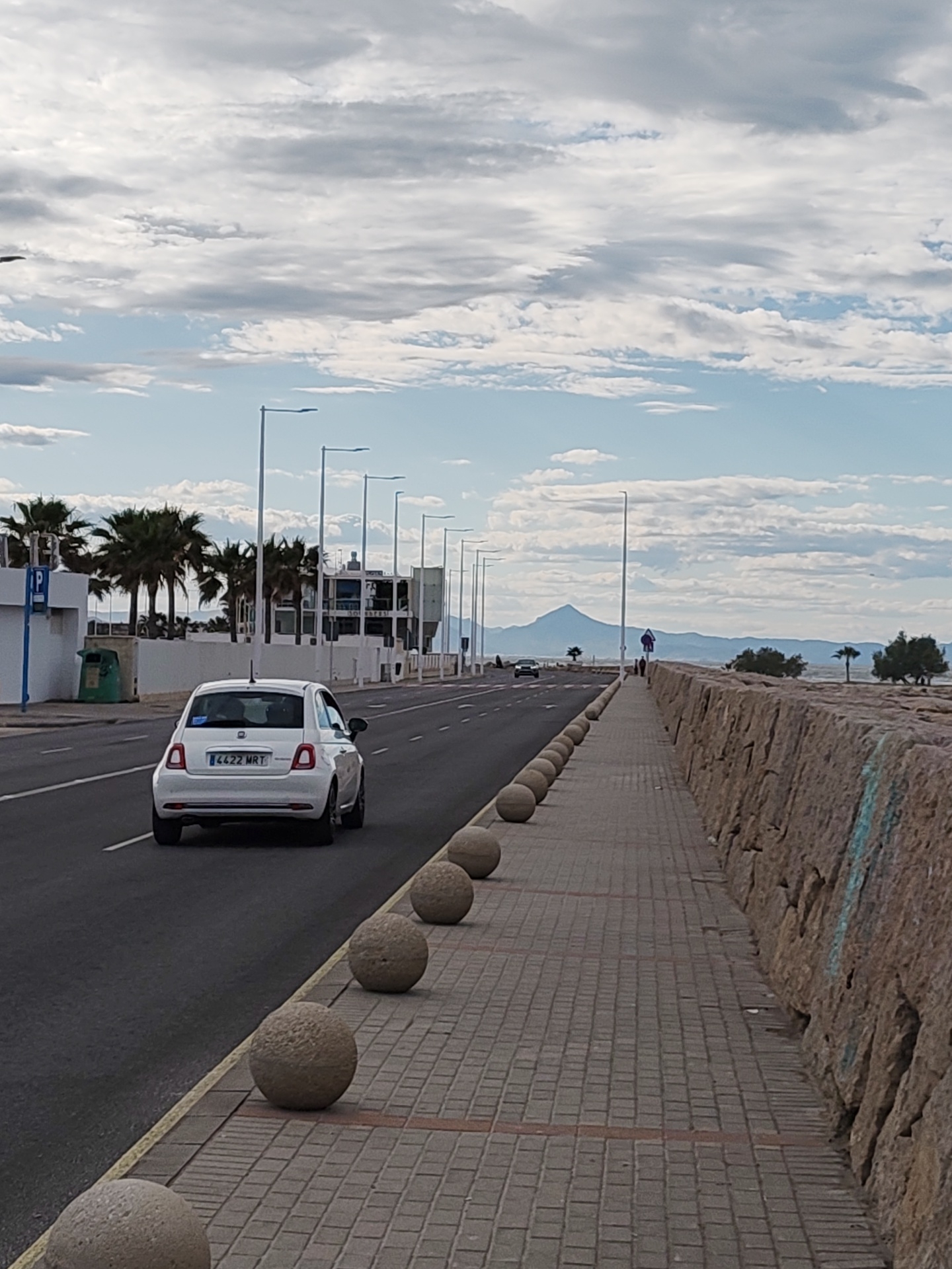The image shows a street scene in Dénia, Spain. A white Fiat 500 with the license plate "4422 MRT" is traveling on the left side of the road. The road is flanked by a row of palm trees on the left and a stone barrier and walkway on the right. Spherical concrete bollards separate the road from the walkway. The sky is partly cloudy. In the distance, a mountain can be seen. A couple of people are walking along the walkway.