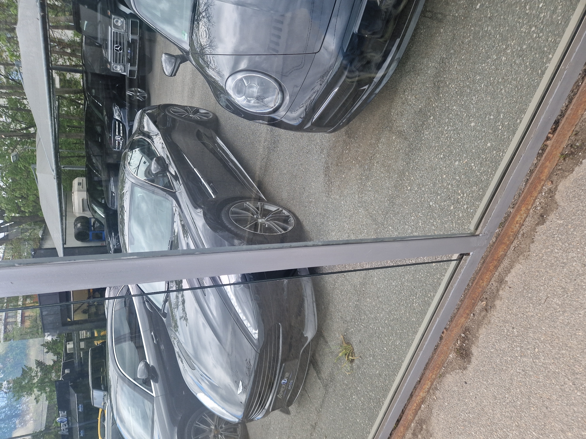 The image shows a reflection in a window, capturing the view of a car dealership with various vehicles. The dealership is located in an urban environment. The cars include a dark-colored Porsche, and Aston Martin, along with several other models, which appear to be Mercedes-Benz. The environment seems to be well-lit, with daylight illuminating the cars and the surrounding street.