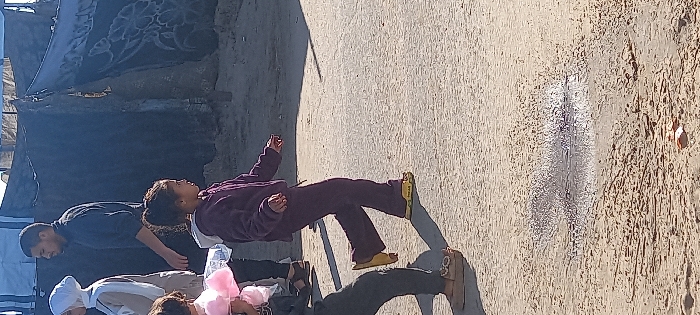 The image depicts an unpaved outdoor area in Khan Yunis, Palestinian Territory, under strong sunlight. In the foreground, a person's lower leg, clad in dark pants and light brown loafers, is visible on the left, holding a pink, fluffy object resembling cotton candy. A young girl, positioned centrally, is captured in mid-stride moving towards the right. She wears a purple fleece tracksuit and yellow clogs, with the word "POLAR" visible on one. Her dark hair is pulled back. Behind her, a woman in a white hijab and a light-colored top walks to the right, followed by a man with short dark hair and a beard, wearing a dark long-sleeved shirt, also proceeding right. The background features several makeshift structures, appearing as tents or shelters, constructed from dark tarpaulins, some displaying patterned designs. A small, reflective puddle of liquid is present on the uneven ground in the lower center.