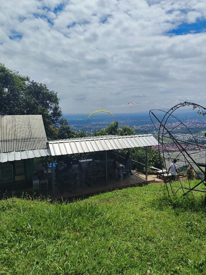 The image shows a view from a hilltop overlooking the city of Majalengka, Indonesia. The coordinates -6.846, 108.204 place the image at the Puncak Gajah, a popular viewpoint in Majalengka. Two paragliders are visible in the sky. A small, corrugated metal building with an open roof, covered by a metal awning, stands on the hillside. A few figures are visible beneath the awning and a single figure is standing on a path in the foreground. There are many trees on the hillside and a metal structure in the foreground. The sky is mostly blue with a few white clouds. The ground is covered in green grass.