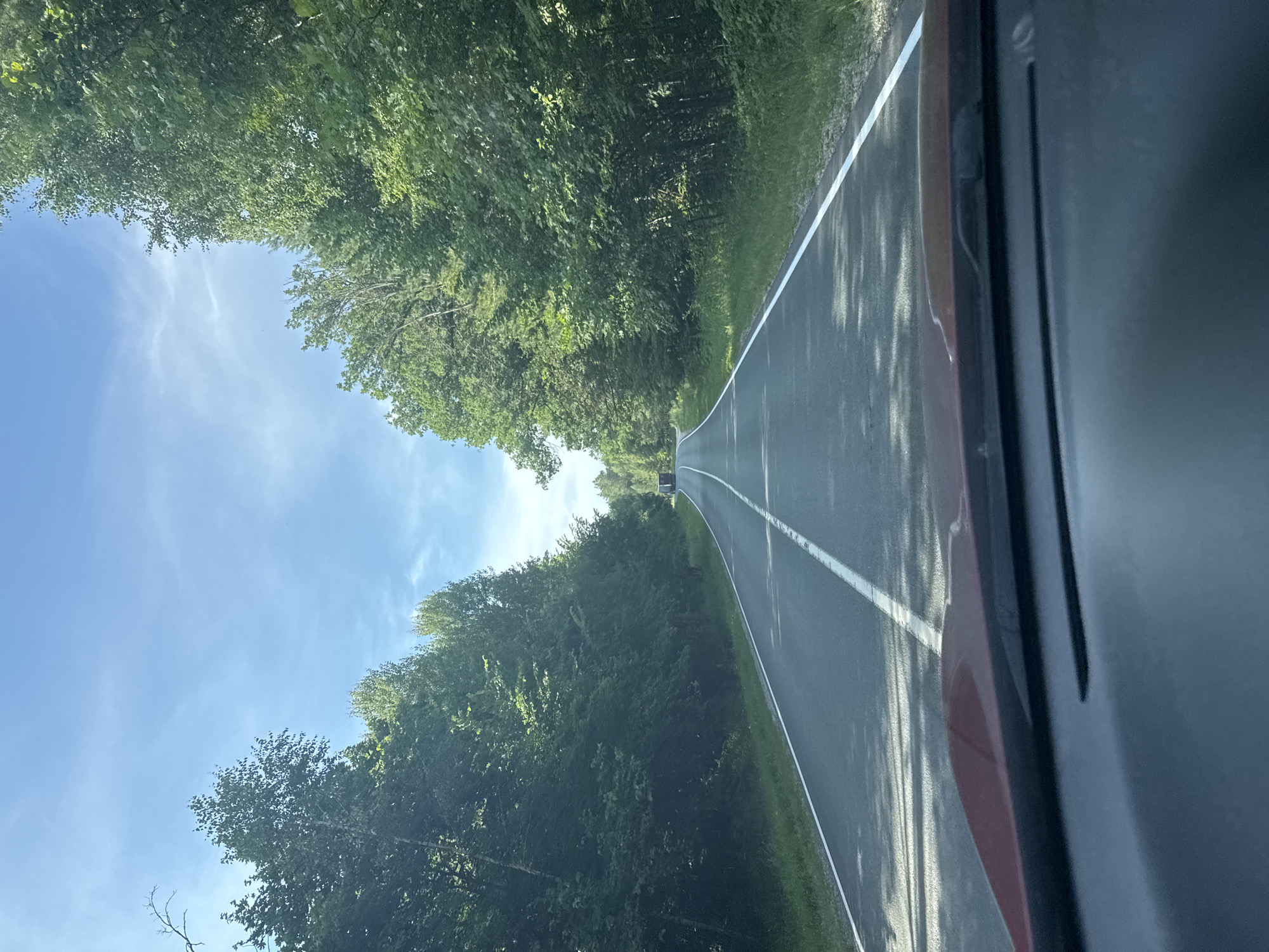 An asphalt road curves through a dense forest under a clear blue sky with scattered clouds. The road is marked with a solid white line on the right edge and a broken white line down the center. Trees line both sides of the road, their green foliage creating a canopy. In the distance, a red truck is visible on the road. The foreground shows the dashboard of a red car. The scene appears to be a sunny day. The location is the city of Ivanovka, Russia.