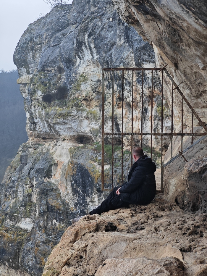 The image captures a solitary male figure sitting on a rocky ledge within a rock-hewn dwelling or monastery cell, typical of the rock monasteries located in the Shumen Plateau Natural Park, near Shumen, Bulgaria. The man, observed from behind and slightly to his right, is wearing a dark, padded winter jacket and dark trousers. He is seated on an uneven, light brown and grey rock surface, facing left and looking outward over a precipice. Directly in front of him, a large, rusted metal grate with vertical bars spans the opening of the rock structure, secured into the multi-toned rock walls. To the man's left, beyond the grate, a monolithic, weathered limestone cliff descends sharply, exhibiting streaks of grey, white, and tan, with small patches of dark vegetation in its crevices. The distant background on the left reveals a blurry outline of a forested landscape under an overcast sky. The overall lighting is diffused, suggesting a cloudy day.