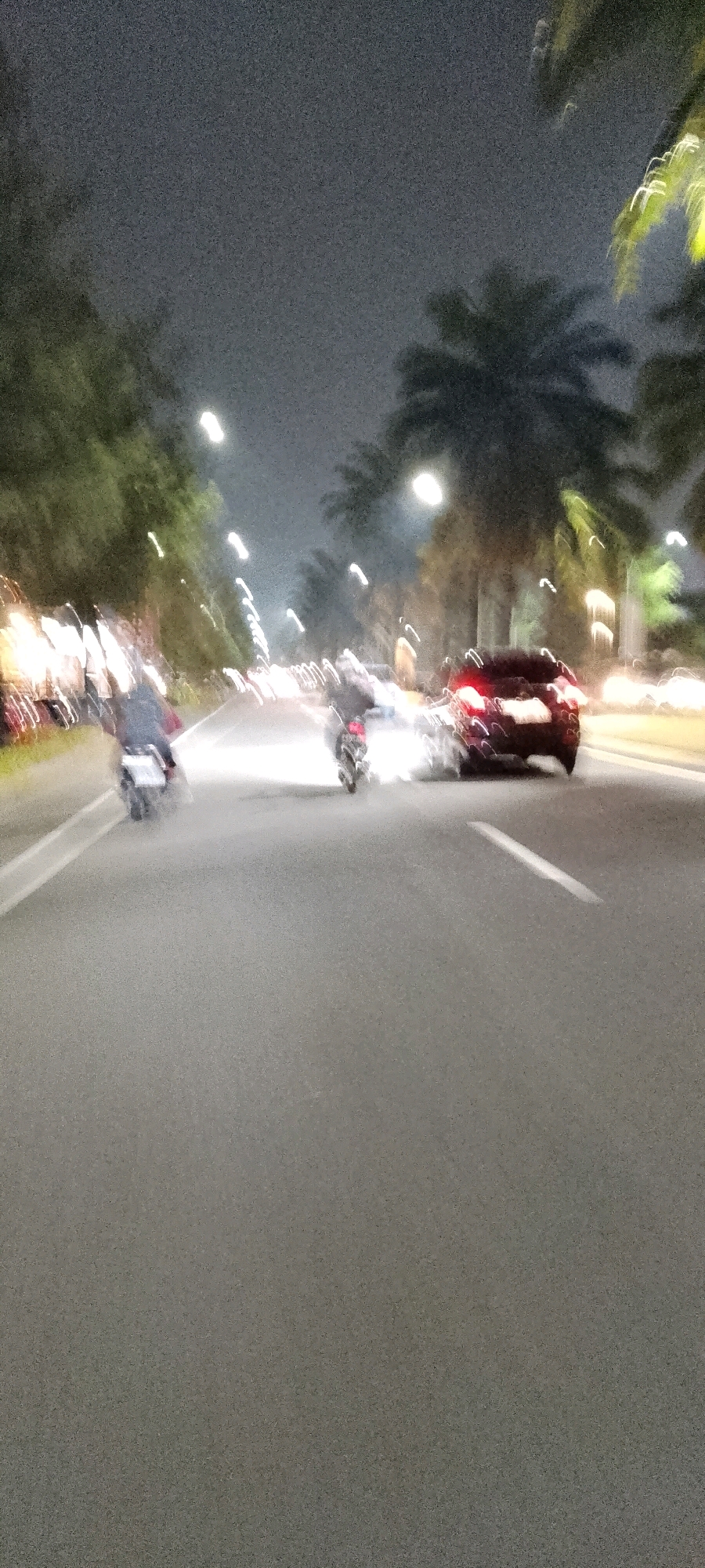 This is a low-light, motion-blurred photograph taken at night on a paved road in Gunung Putri, Indonesia. The scene depicts a traffic situation with several vehicles. A dark-colored car, possibly a sedan, is visible in the right lane, its brake lights illuminated. To the left of the car, a motorcycle is also present, with its rider blurred due to motion. Further to the left and ahead, another motorcycle is visible, also blurred. The road has white lane markings. Along the sides of the road, trees and streetlights are visible, contributing to the blurred streaks of light characteristic of night photography with motion. The sky is dark and grainy.