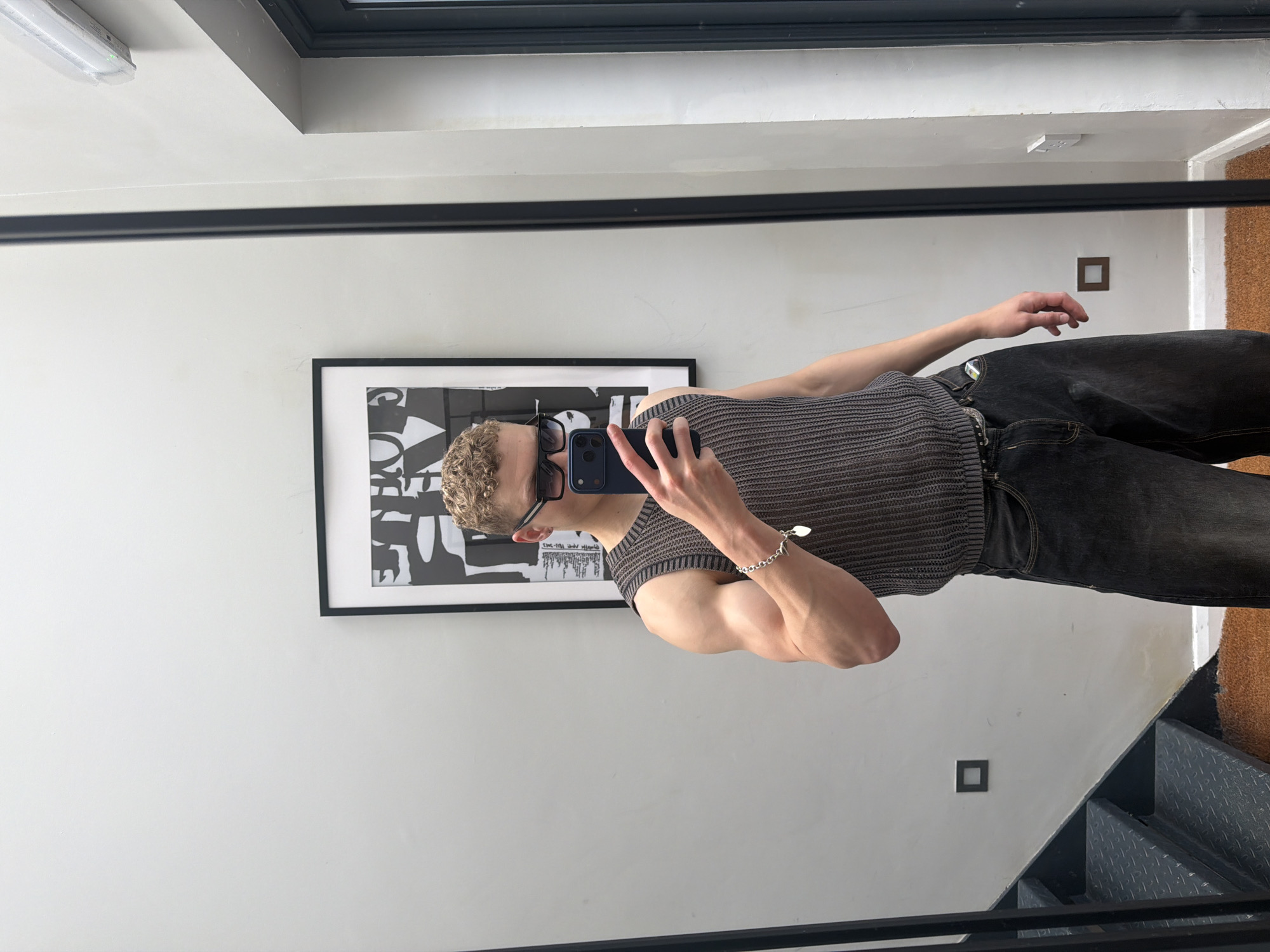 A fair-skinned person, possibly male, is taking a selfie in a mirror. The person has short curly blonde hair and is wearing a gray sleeveless knit shirt, dark-wash jeans, and sunglasses pushed up on their head. A silver chain bracelet is visible on their left wrist. The phone is dark blue.
The background includes a white wall with a black-framed piece of abstract art hanging on it. There is a square electrical outlet on the wall. Above the person's head is a white ceiling with a fluorescent light fixture and a window frame. To the right of the person are metal stairs and a cork wall. The photo is taken in a building. The location is not identifiable as being in the city of London.