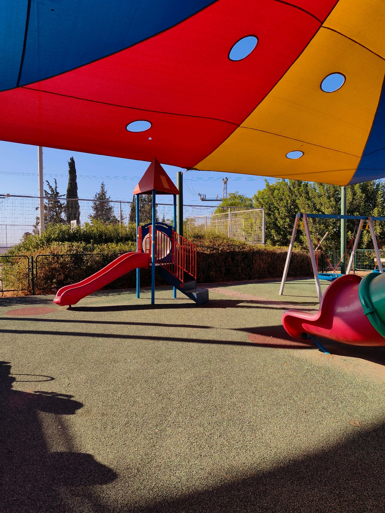 The image depicts a children's playground in the city of Lachish Regional Council, Israel. The playground features a red slide, a swing set, and a tubular slide in pink and green. A colorful canopy, divided into blue, red, and yellow sections, provides shade over the play area. The ground is covered with a textured, green surface. In the background, a fence encloses the playground, with trees and vegetation visible beyond it. The overall scene suggests a bright, sunny day, with shadows cast by the play structures and canopy. There are no people present in the image.