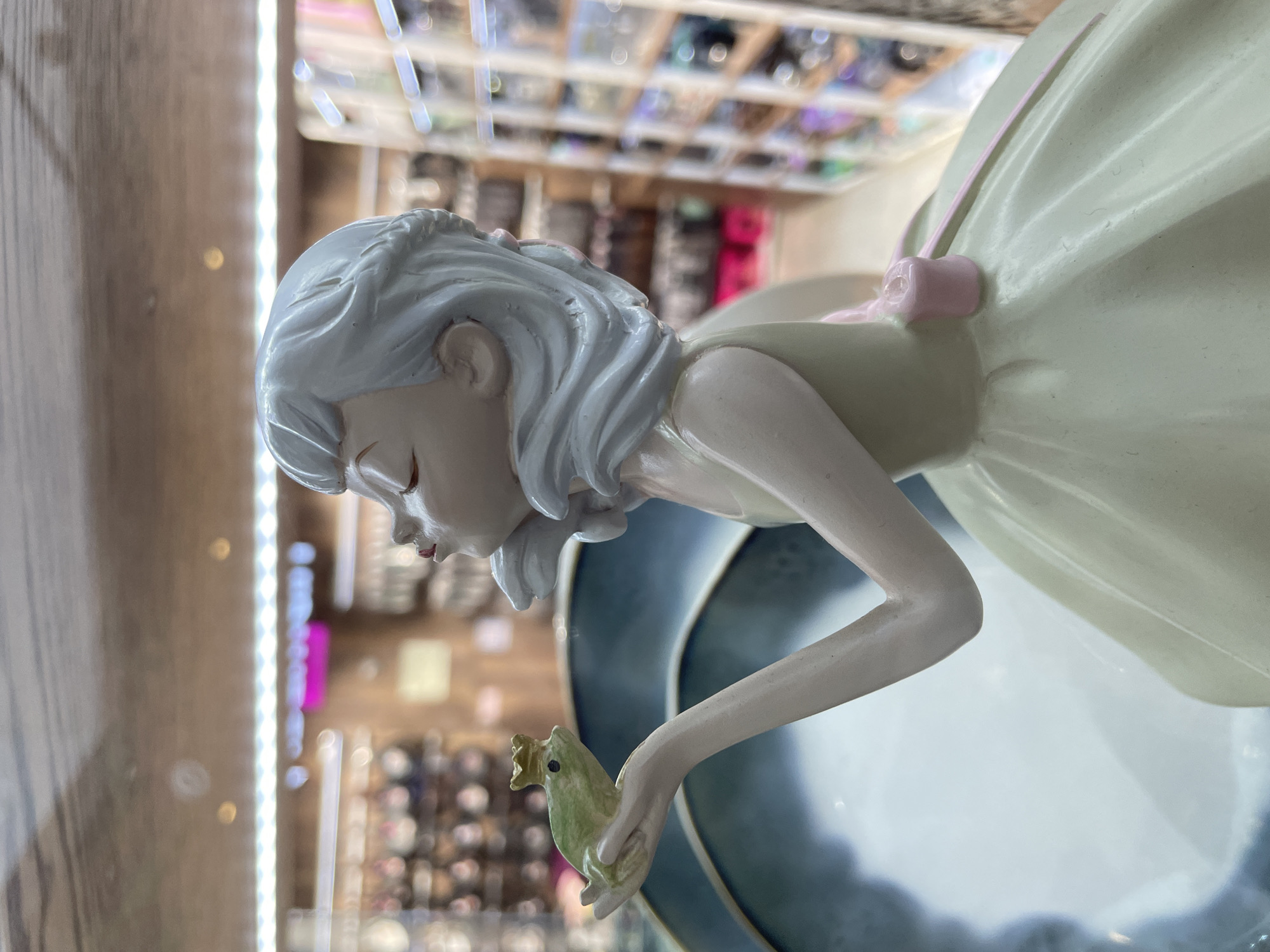 A porcelain figurine of a girl holding a frog is displayed in a glass cabinet. The girl has white hair and a pale yellow dress with a pink bow. She is looking down at the green frog she holds in her hand. The background is blurred, but suggests a retail environment with rows of eyewear on display. The location is the city of Kommunarka, Russia.