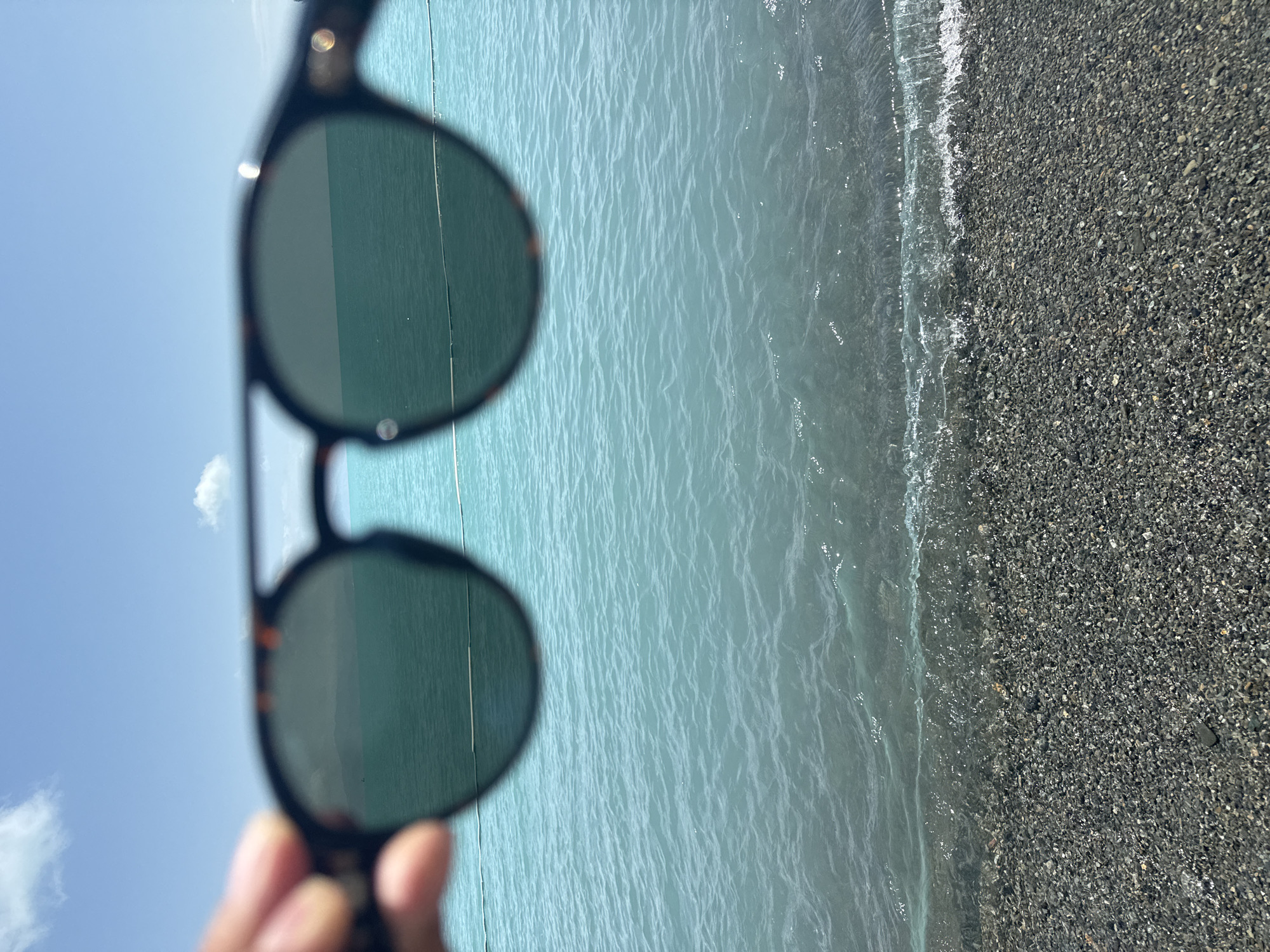 The image shows a person holding sunglasses in the foreground, obscuring a portion of the view. Beyond the sunglasses, a body of turquoise water stretches towards a distant, hazy shoreline with mountains in the background. The water is calm with small ripples. A narrow band of dark, pebble-strewn beach is visible at the bottom of the frame, with gentle waves lapping at the shore. The sky above is bright blue with a few scattered white clouds. The scene appears to be a sunny day at a lakeside or seaside location. The location is identified as Tsamakaberd, Armenia, suggesting a view of Lake Sevan.