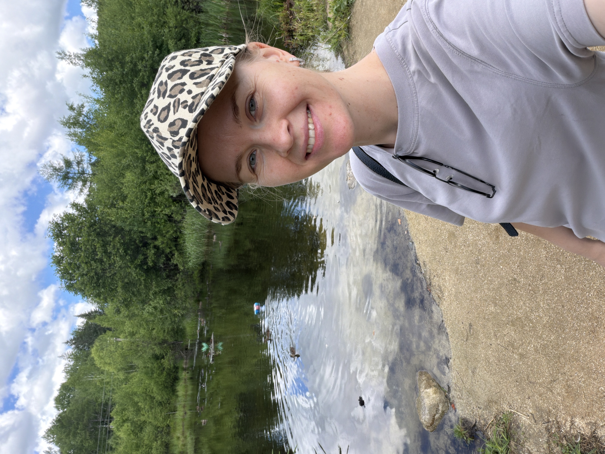 A woman is taking a selfie in front of a lake on a cloudy day. She is wearing a grey t-shirt and a leopard-print baseball cap. She is smiling at the camera. There are trees and foliage surrounding the lake, with some ducks swimming in the water. The woman is standing on a sandy shore.