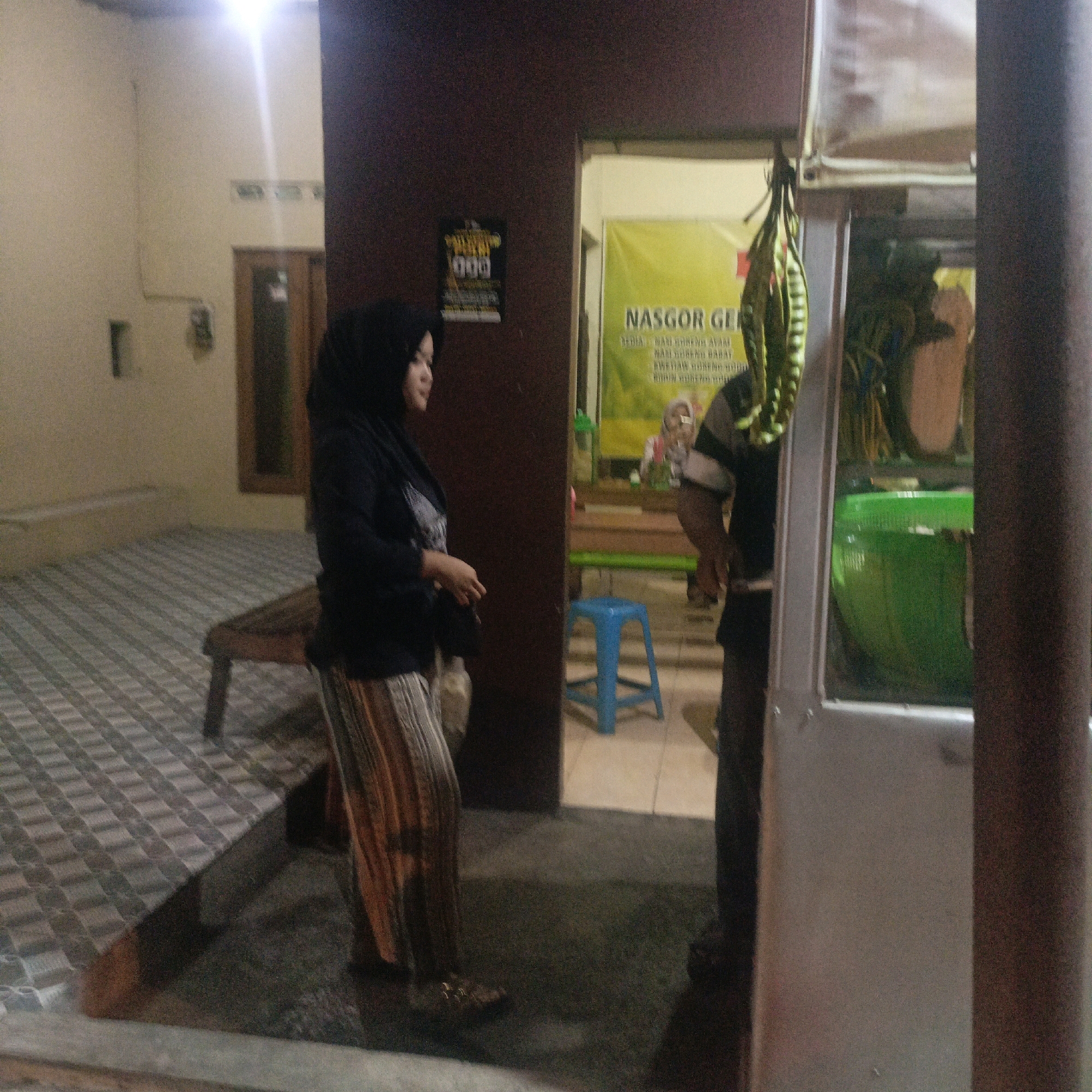 The image captures a nighttime scene at what appears to be a food stall in Salatiga, Indonesia, specifically near the intersection of Jalan Diponegoro and Jalan Kartini, a known area with street food vendors. In the foreground, a woman wearing a black hijab and a patterned skirt stands to the left, facing away from the camera and towards an open doorway. She is wearing sandals. Behind her, a bench is visible.
The doorway leads into a brightly lit interior where a yellow banner with Indonesian text, possibly advertising a food item like "NASGOR" (Nasi Goreng or fried rice), is prominently displayed. Inside, a person wearing a striped shirt, partially obscured, is seen working at the stall. A blue plastic stool is placed just inside the doorway.
To the right of the doorway, a food cart or stall is visible, with its metallic exterior reflecting light. A large green container is prominent within the cart, and what appears to be dried vegetables are hanging from a string above it. The overall environment suggests a bustling street food setting.
