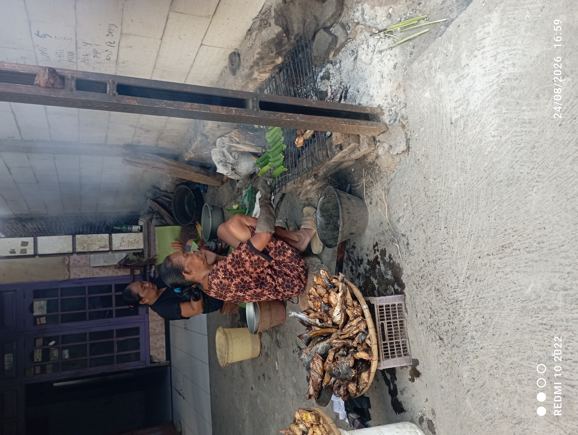 In Karangsari, Indonesia, two women are engaged in food preparation outdoors. One woman, wearing a dark shirt, sits to the left, while another woman in a patterned dress sits to the right, closer to the cooking area. In front of the seated women, a woven basket overflows with dried fish, suggesting they are preparing them for sale or consumption. To the right, a makeshift grill, constructed with metal bars and brick, holds several pieces of food cooking over hot embers. Green vegetables, possibly corn or bananas, are arranged on the grill. The environment appears to be a simple outdoor workspace, with concrete flooring and a partially visible building in the background. The scene is marked by smoke rising from the grill, indicating active cooking. A bucket and other containers are scattered around the area, typical of an outdoor market or street food vendor setting. The timestamp on the image indicates it was taken on August 24, 2026, at 4:59 PM.